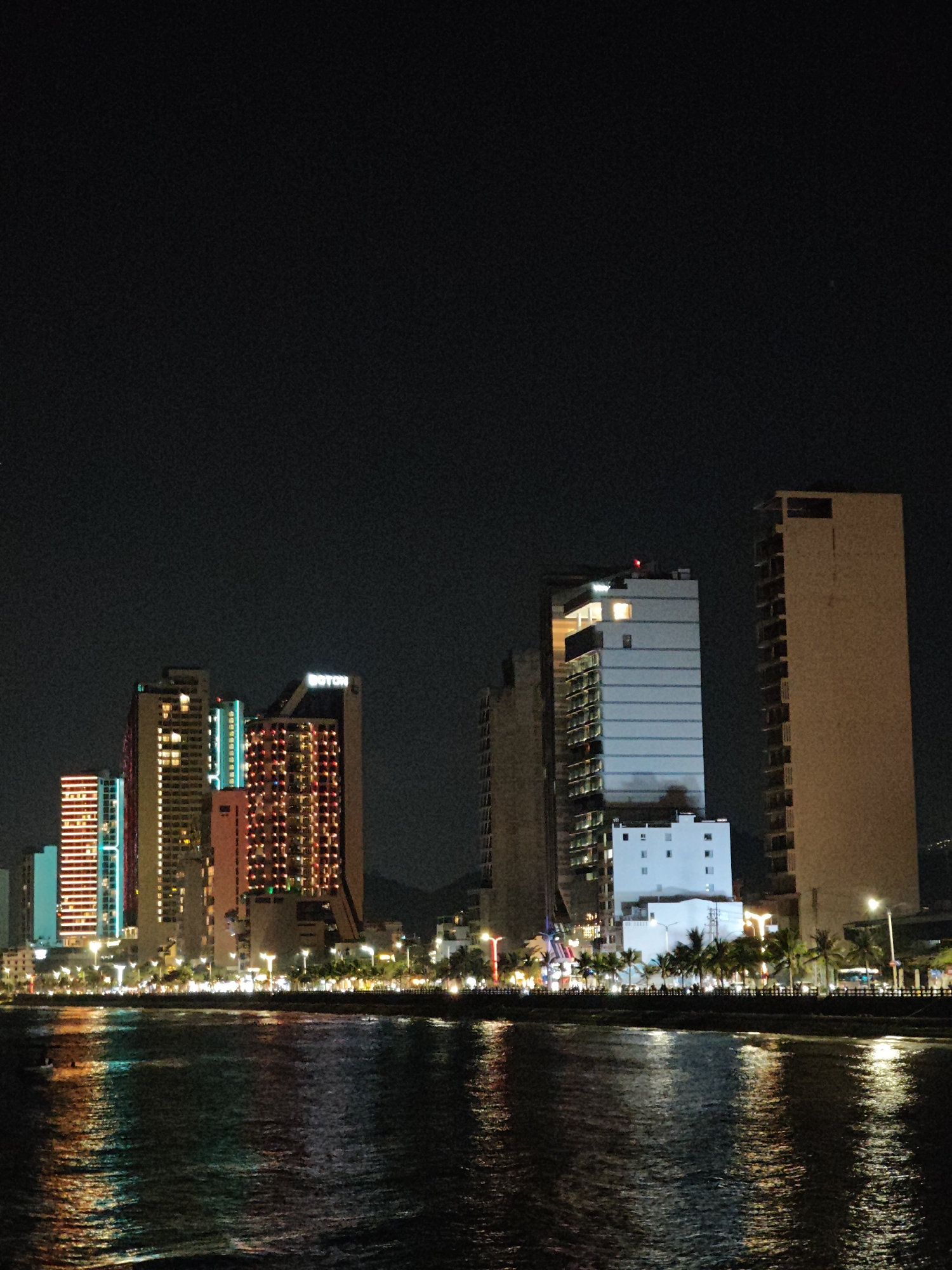 The image depicts a nighttime cityscape, featuring a row of illuminated buildings along a waterfront. The buildings vary in height and architectural style, with some displaying brightly colored lights. A body of water, likely a sea or ocean, reflects the city lights, creating a shimmering effect. The sky is dark, providing a stark contrast to the illuminated city. A row of palm trees lines the waterfront, further defining the urban landscape.