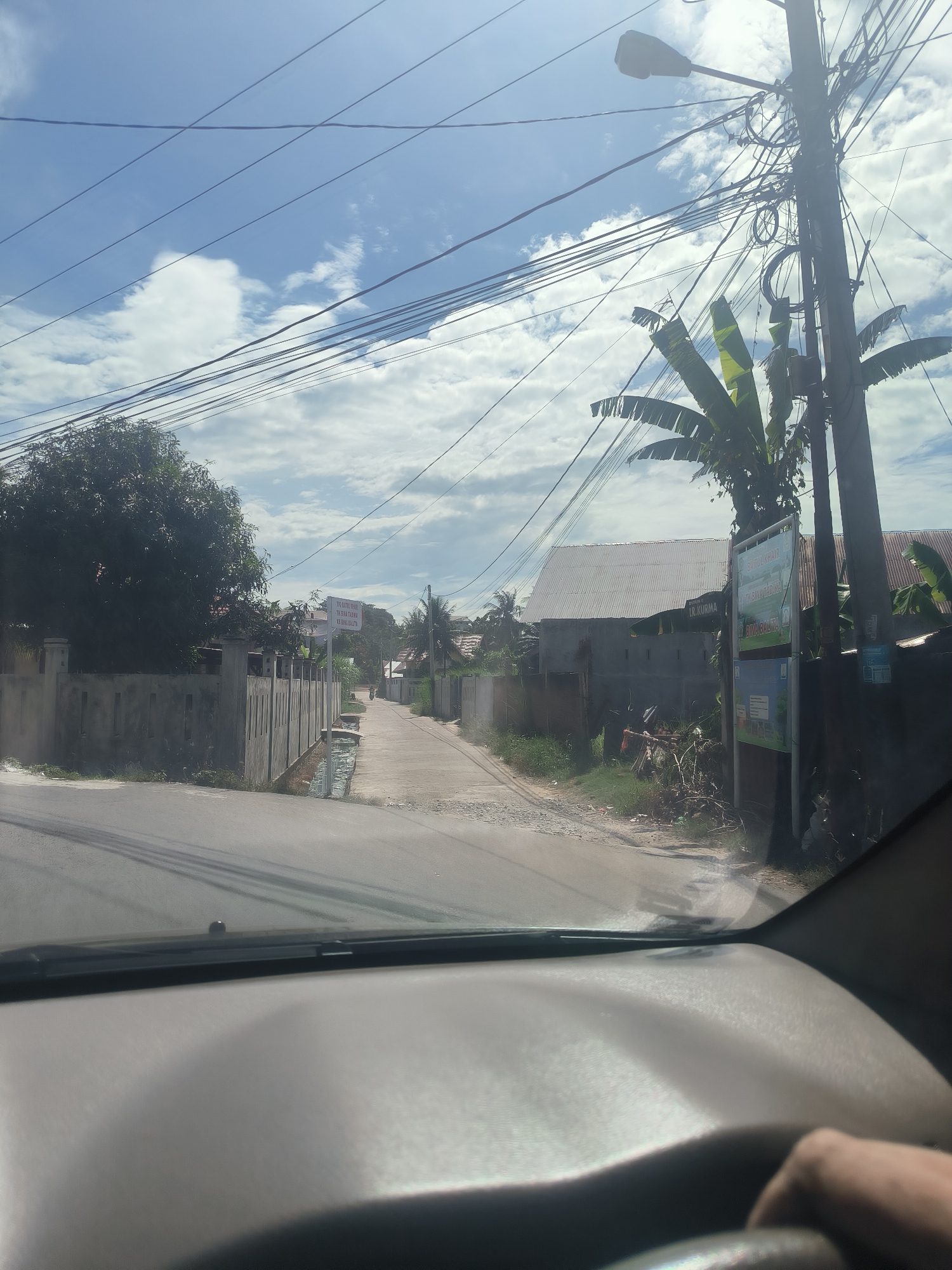 The image presents a street view from inside a vehicle in Lhokseumawe, Indonesia, on a bright, sunny day with a clear blue sky and scattered white clouds. The foreground shows the car's dashboard and the top portion of a steering wheel, with a hand visible on the right.
The asphalt road ahead appears somewhat worn and stretches into the distance, with a slight curve to the left. On either side, concrete fences separate properties from the road. The landscape features abundant tropical vegetation, including large trees and prominent banana palms, especially near the utility pole on the right.
A large electrical pole stands prominently on the right, burdened with a dense network of overhead wires that crisscross the sky. A street light is also attached to this pole. Visible signs provide local information:
* A white sign on the left side of the road reads "THE BATEL DALL", "TK BINA TADINA", and "KABINA BALITY".
* A larger, multi-panel sign on the right, near the electrical pole, displays "TUL KHAIR", "TABINATEDING", "LR. KURMA", and "AWABALIE".
The scene depicts a typical residential or semi-urban street, characterized by its utility infrastructure, tropical flora, and local signage under bright daylight conditions.