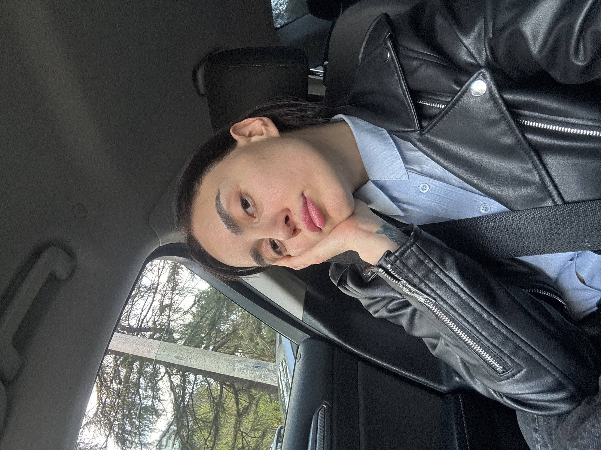 A person is visible in the interior of a vehicle. The person, facing the camera, rests their chin on their hand, wearing a light blue button-down shirt and a black leather jacket, secured by a seatbelt. Visible outside the window are trees, suggesting an outdoor environment. Based on the reflections in the vehicle windows, the surrounding environment has trees in the city of Tbilisi, Georgia.