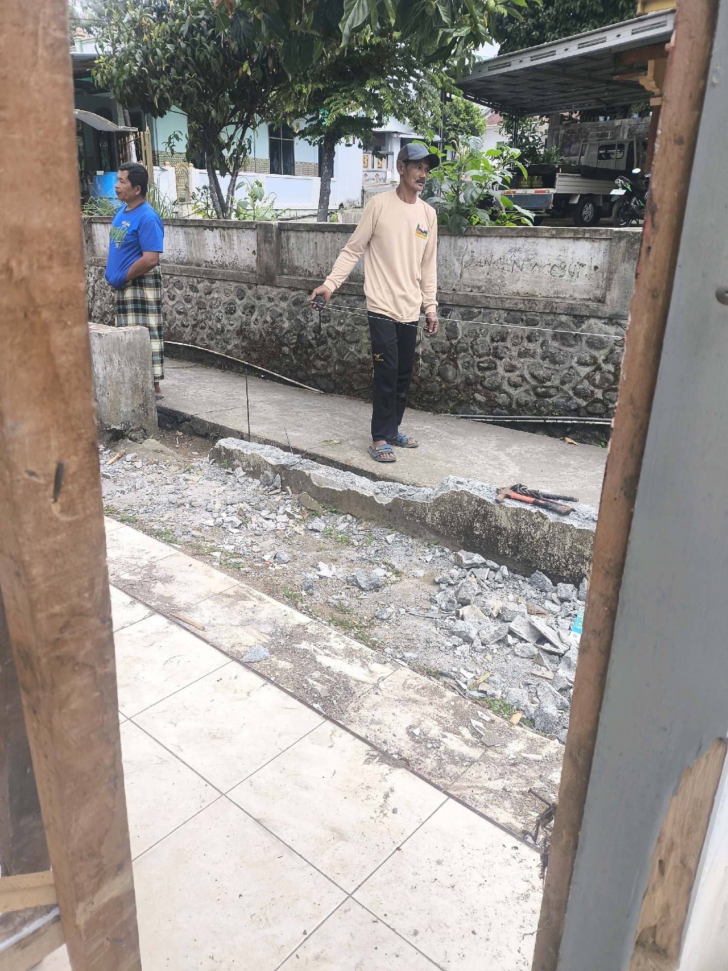 The image depicts a street scene in Senduro, Indonesia. Two adult males are visible. The man on the left, wearing a blue t-shirt and a checkered sarong, stands facing left, looking away from the camera. The man on the right, wearing a light tan long-sleeved shirt, dark pants, and a blue cap, is facing left and appears to be holding a wire or string with his right hand. He is standing on a concrete path next to a damaged concrete structure. Several construction tools, including what appear to be pipe wrenches, are lying on the damaged concrete. The background shows a wall made of rough stones and a covered structure with a vehicle partially visible beneath it. The ground is covered in loose gravel and debris, indicating ongoing or recent construction or repair work.