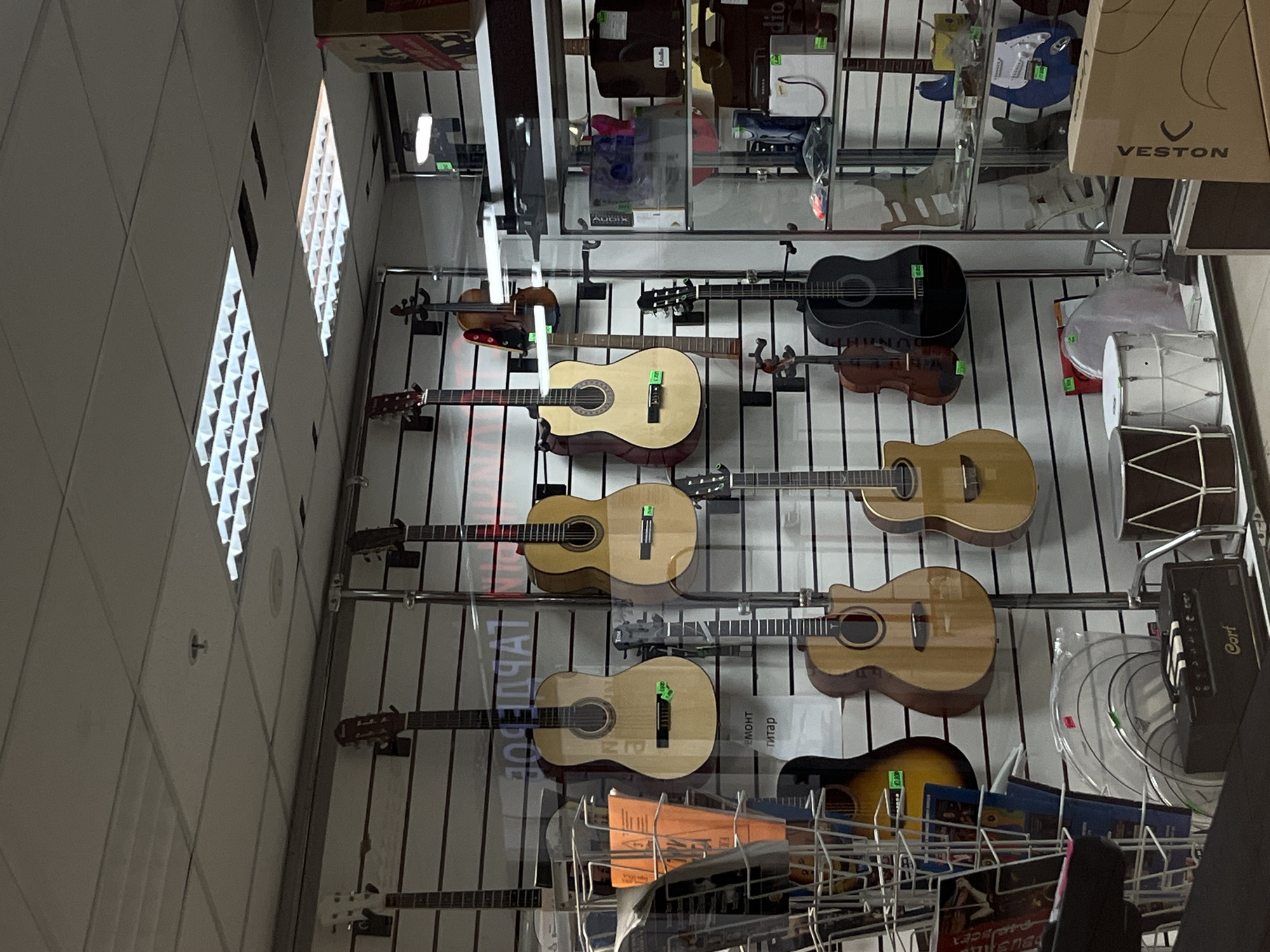 This image shows a collection of guitars displayed on a wall in a store in Klin, Russia. The guitars are of various types and colors. There are drums and other accessories also on display. The image is taken from a low angle, looking up at the wall of guitars. The lighting in the store is artificial.