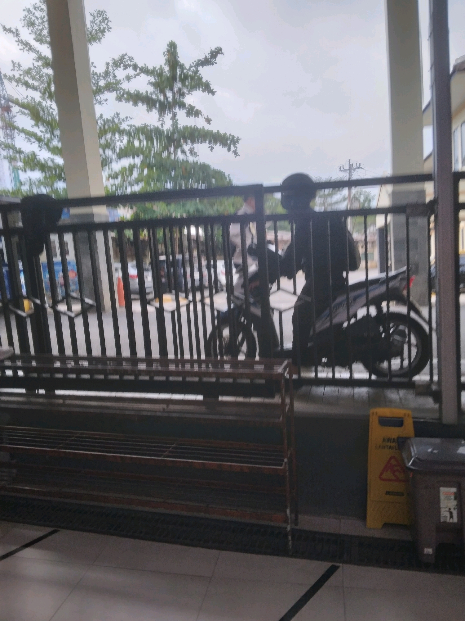 The image depicts an outdoor scene, likely in a public area with a covered walkway or building overhang. In the foreground, there is a multi-tiered metal shelf, possibly for shoes or storage. Beyond this shelf is a black metal fence. To the right of the fence, a yellow cautionary sign with a red triangle and exclamation mark is visible, along with a brown trash bin.
Through the fence, a person wearing a dark helmet and dark clothing is seated on a motorcycle. The motorcycle is light-colored with blue and white accents. The background shows some greenery from trees, a building structure, and a cloudy sky. There are also hints of other vehicles and possibly some construction equipment in the distance. The overall impression is of a somewhat overcast day. The location is identified as Desa Pamijen, Kecamatan Sokaraja, Indonesia.