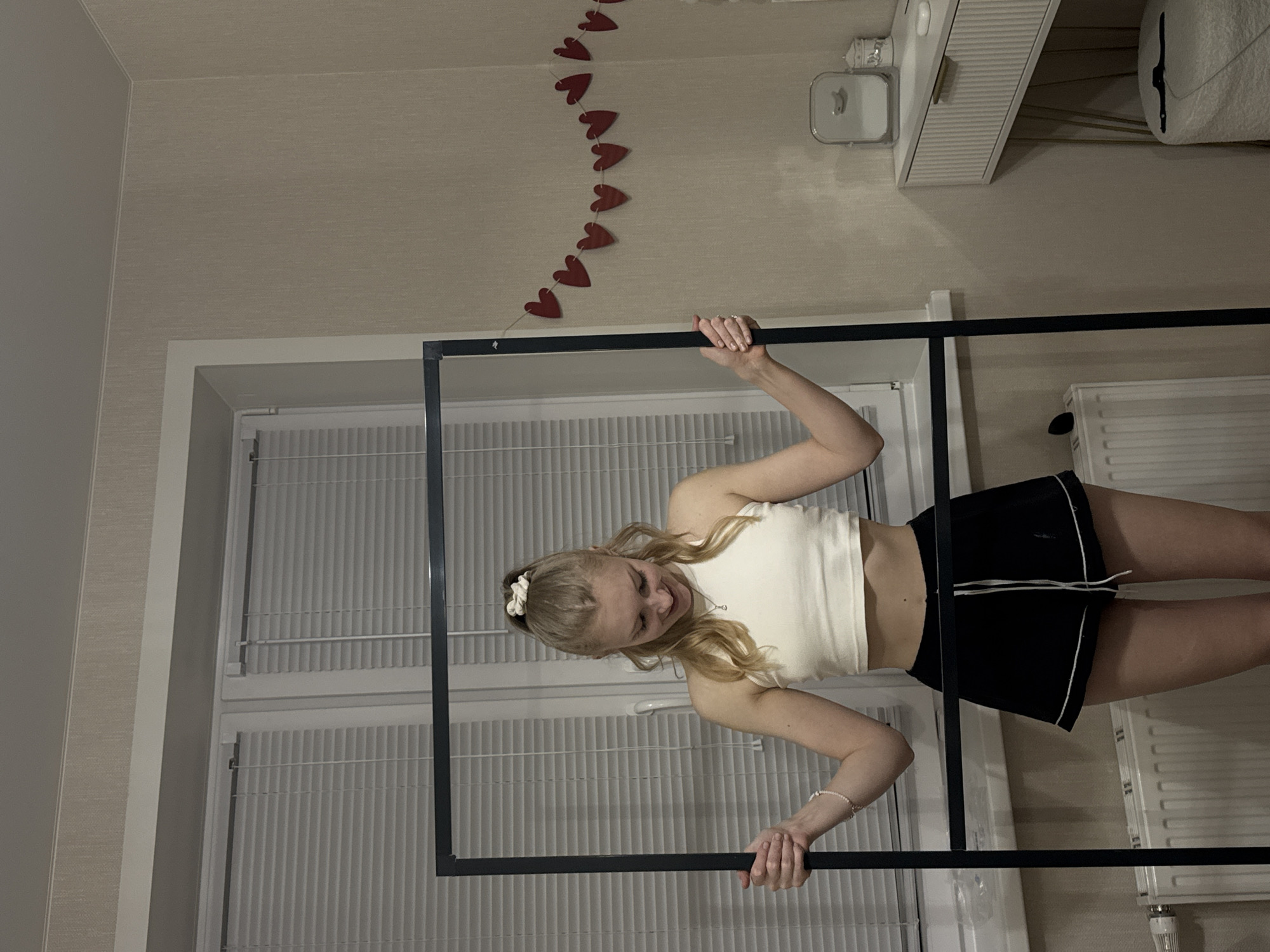 A woman with blonde hair and wearing a white top and black shorts holds a black frame in front of a window. Red heart-shaped decorations hang above the frame. The location is a room with a radiator and furniture. The city and country are not discernible.