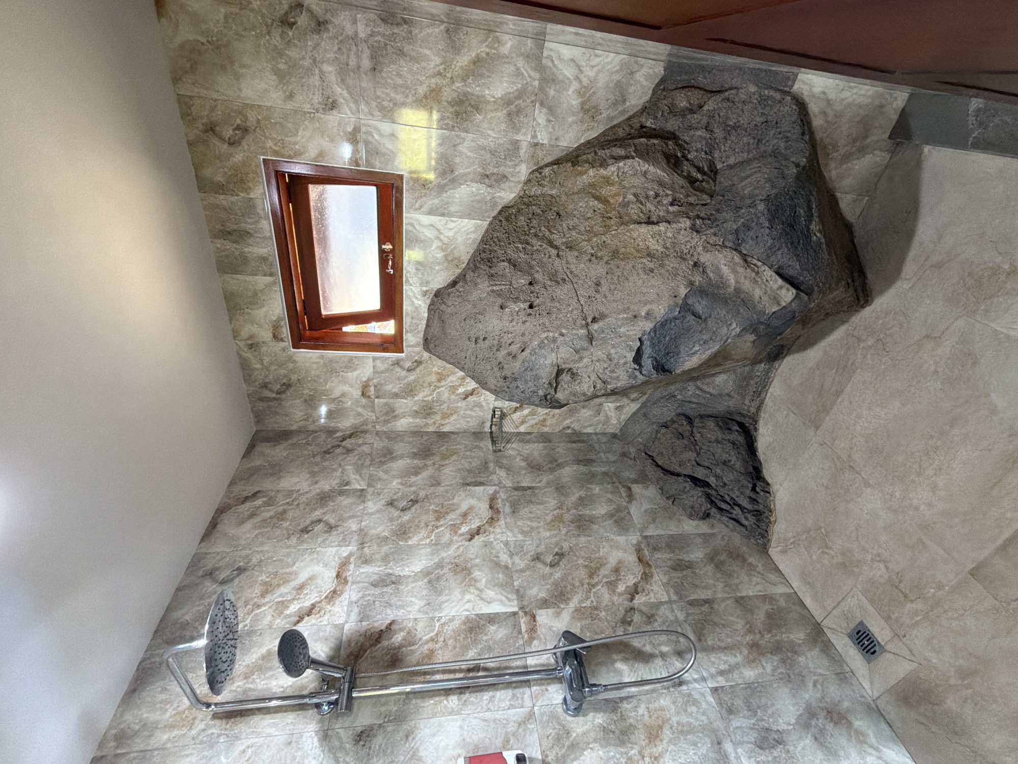 The image shows a unique shower stall with natural rock formations integrated into its design. A large, rough-textured rock is prominently positioned in the corner, with a smaller rock formation adjacent to it. The walls are tiled with a marble-like pattern in shades of beige and brown. A modern chrome shower fixture with two showerheads is mounted on the left wall. The floor is tiled with large, light-colored tiles, and a drain is visible in the foreground. A small, wooden-framed window with frosted glass is located high on the back wall. The overall impression is one of a rustic and naturalistic bathroom design. The location is identified as Abang, Indonesia.