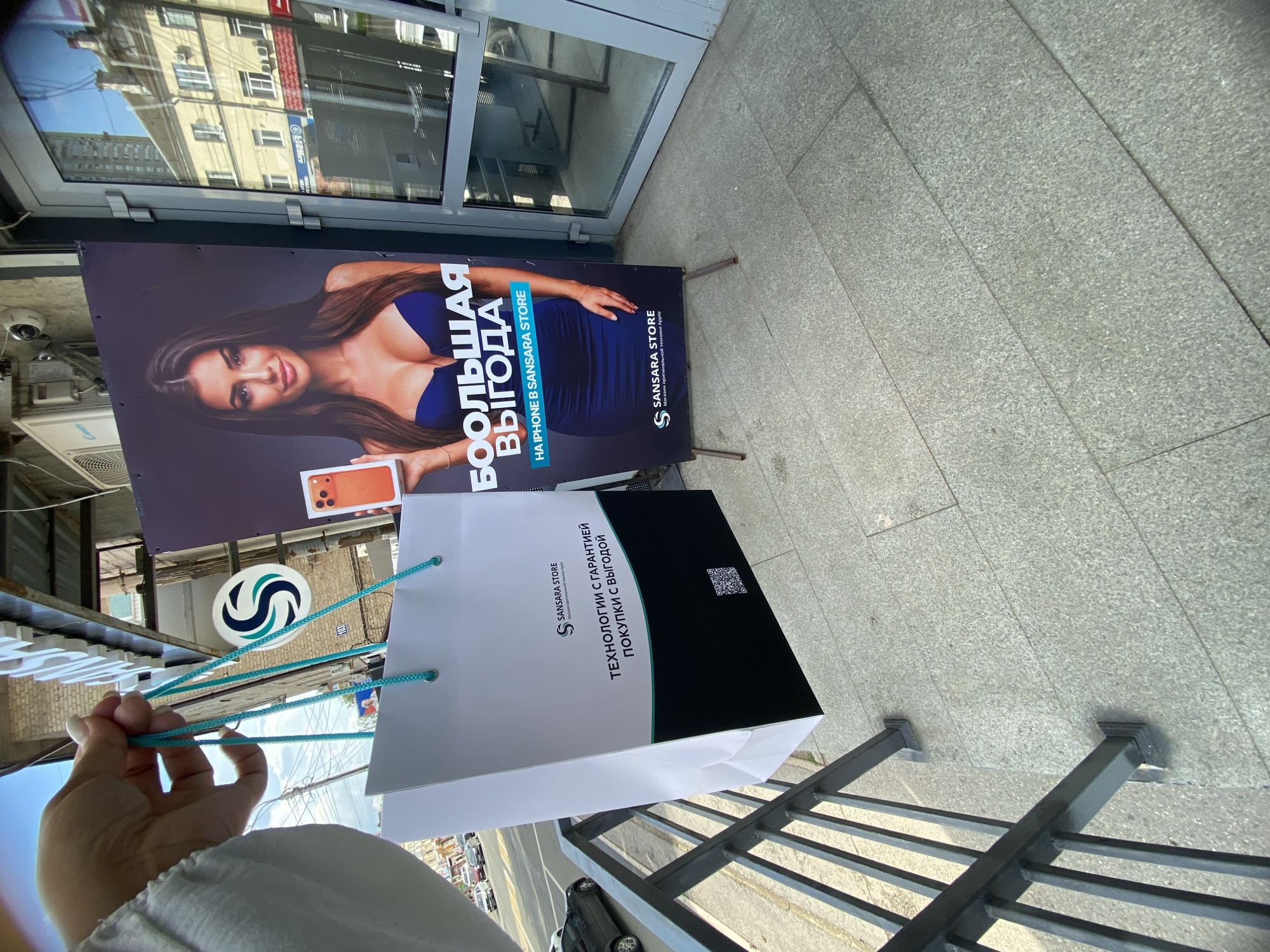 In Ufa, Russia, a person's hand holding a white paper shopping bag with teal handles is pictured. The bag features the "SANSARA STORE" logo and text in Russian indicating "technologies with a guarantee, purchases with profit." A QR code is also visible on the bag. To the right of the bag, a large advertisement displays a woman holding an orange iPhone and the Russian phrase "BIG PROFIT ON IPHONE IN SANSARA STORE." The store's logo is also present on the advertisement. The background shows a building with windows and air conditioning units, and a street with cars. A metal railing is in the foreground.
