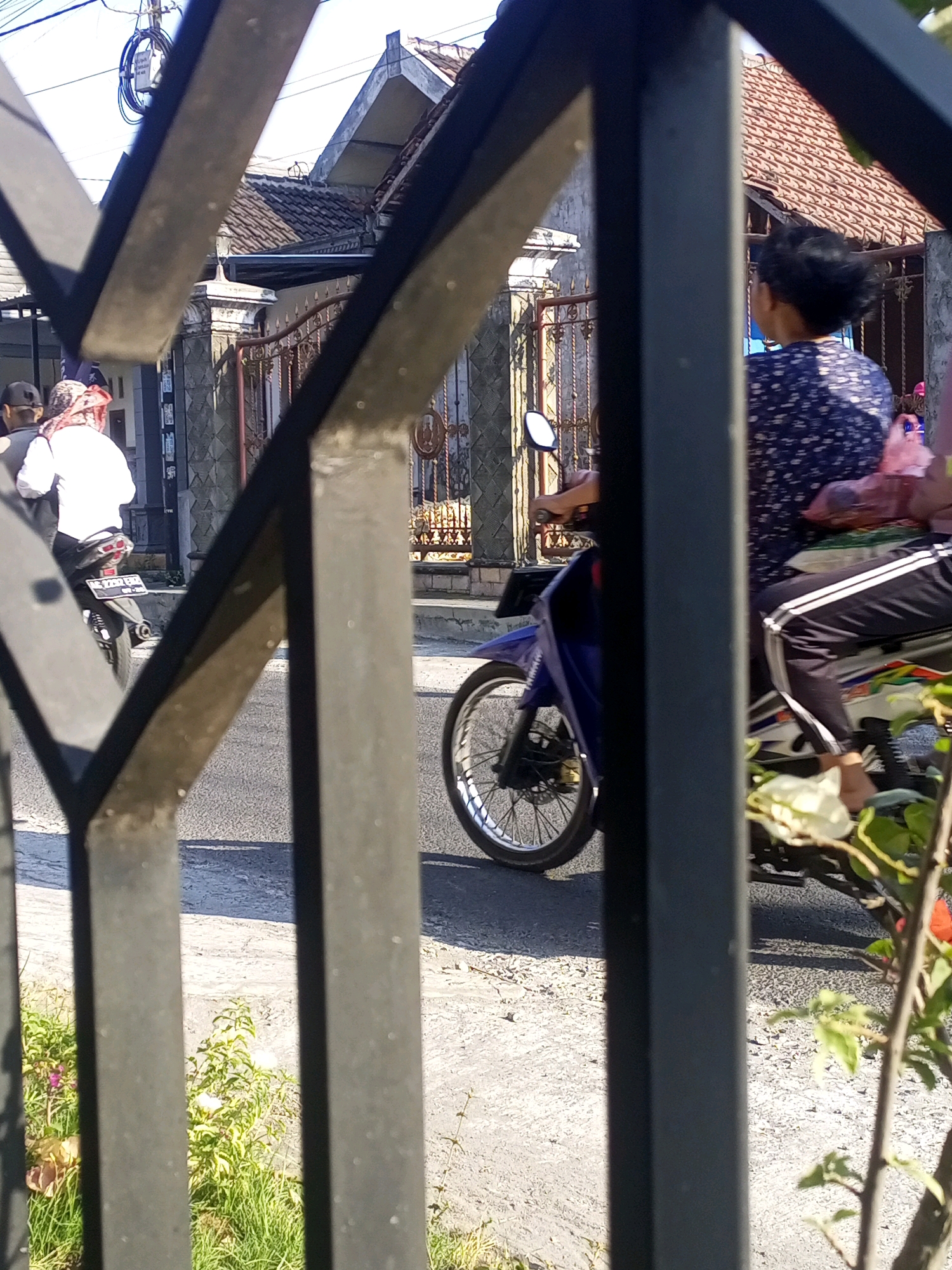 In Ngasem, Indonesia, two individuals are seen on a street, with a black metal fence in the foreground obscuring parts of the scene. The person in the front is wearing a white shirt and a patterned headscarf, riding on the back of a motorcycle. The motorcycle has a white license plate with black lettering. Behind them, another person is riding a blue and white motorcycle, wearing a dark blue floral patterned shirt and black pants with white stripes. They appear to be carrying a pink plastic bag on the back of their motorcycle. The background shows a residential area with houses and a gate. The lighting suggests it is daytime.