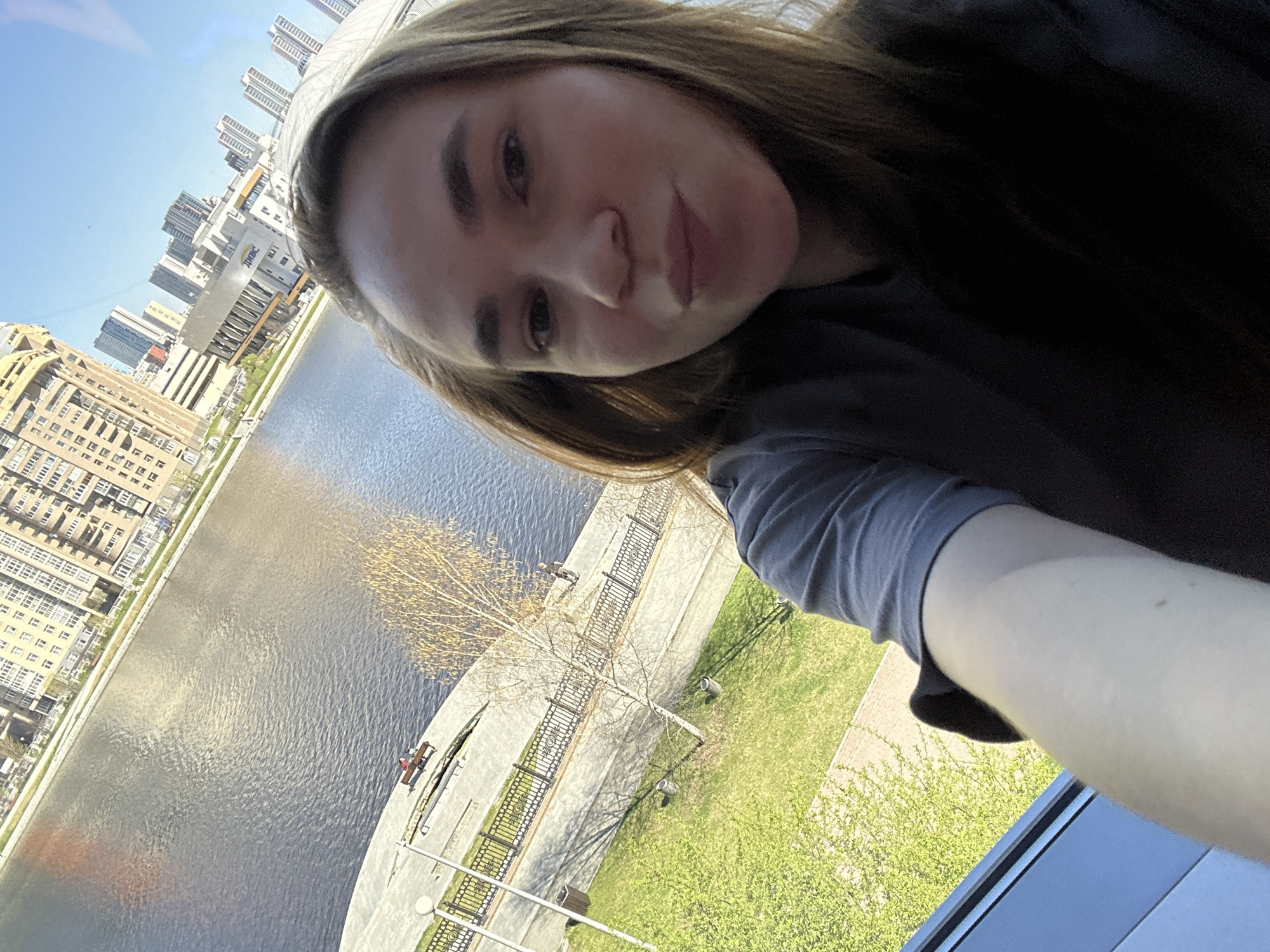 A selfie of a fair-skinned woman with brown hair, wearing a dark top with a gray sleeve, is framed against a cityscape. In the background, a river flows between buildings in what appears to be an urban environment. A walkway and park area with trees and streetlights line the river. People sit on benches along the riverfront. The location is Yekaterinburg, Russia, as indicated by the "ЕльцИн ЦЕНТР" sign on a building visible in the background.