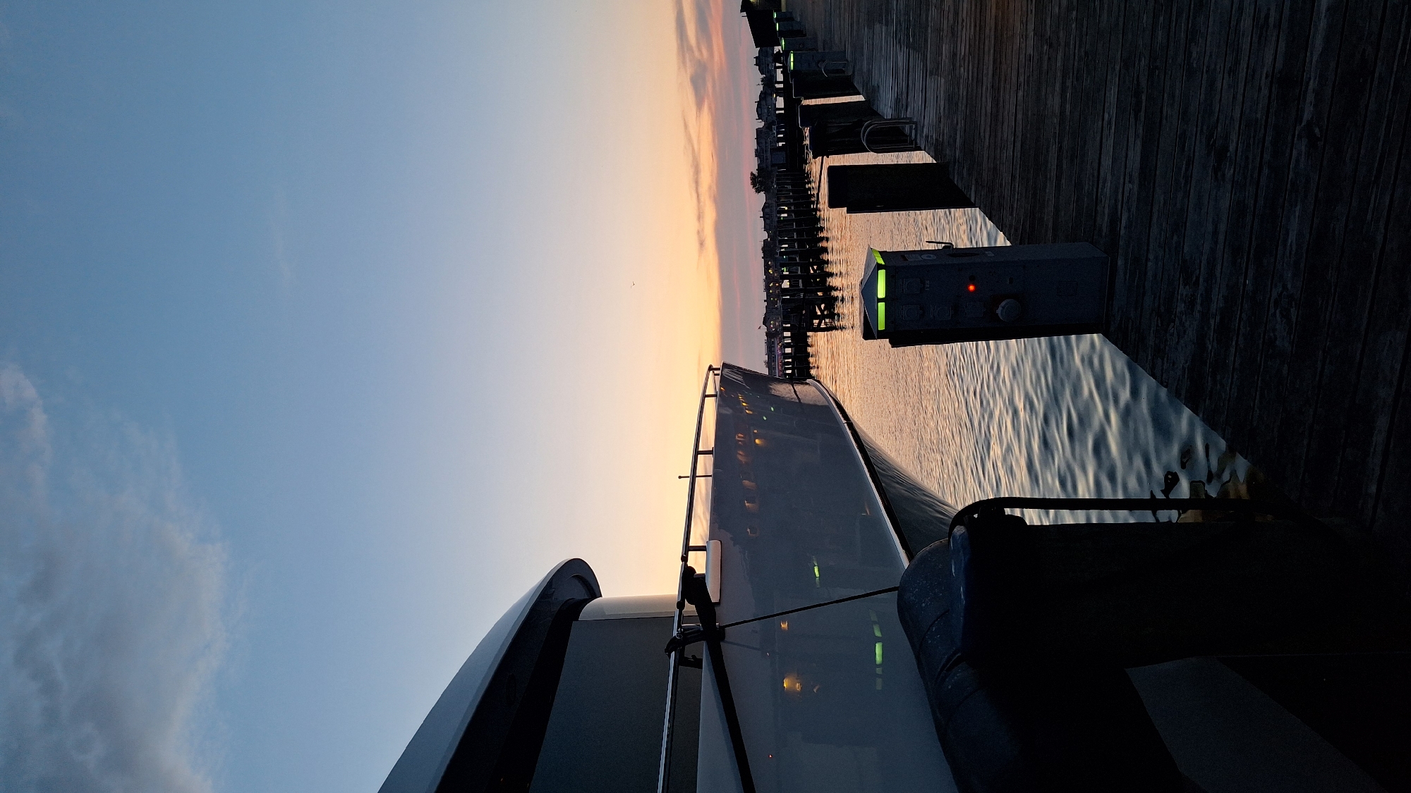 The image depicts a marina in Nantucket, United States of America, during the twilight hours. A large, white yacht is docked on the left, its sleek hull reflecting the soft light of the setting sun. The yacht is tied to a wooden pier that extends into the water on the right. The pier is lined with mooring posts, some of which have green lights on top and a red indicator light on a control box. The water is calm, with gentle ripples reflecting the sky and the yacht. In the distance, a row of buildings and a dock with boats can be seen. The sky is a gradient of soft blues and oranges, with a few wispy clouds. No people are visible in the scene. The overall atmosphere is one of tranquility and serene beauty.