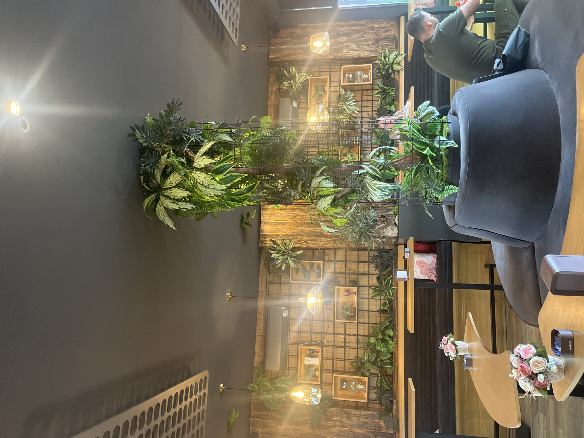 A daytime interior shot reveals a cafe or restaurant setting. A man in an olive green shirt sits in a dark grey chair in the upper right of the frame, facing towards the center. To the left, a vertical metal grid adorned with lush green plants extends from the left wall, reaching towards the ceiling. Further back, a wooden wall serves as a backdrop, decorated with lights and small framed items amidst more greenery. In the foreground, there are wooden tables with small flower bouquets. The location is not explicitly mentioned in the image but based on the request, it can be inferred to be in Plovdiv, Bulgaria.