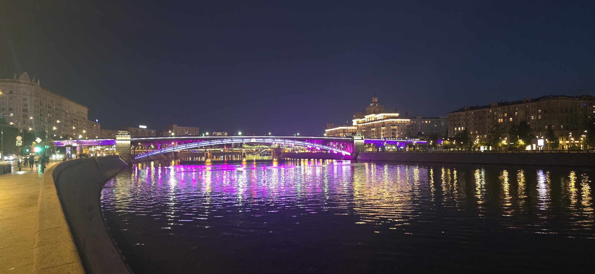 This is a nighttime panorama of a city river with a bridge illuminated by purple and white lights. On the left bank, there is a wide pedestrian walkway with streetlights and several people walking and cycling. Beyond the walkway, there are buildings with illuminated windows. The bridge is the central focus of the image, arching over the water and adorned with bright lights. The right bank of the river features more buildings, also lit up, and trees lining the embankment. The water reflects the colorful lights from the bridge and the surrounding city, creating a shimmering effect. The sky is dark, indicating it is late at night or early morning. Based on the architectural style of the buildings, the illuminated bridge, and the presence of the Moskva River, this scene is likely located in Moscow, Russia, possibly depicting the Krymsky Bridge or a similar structure.