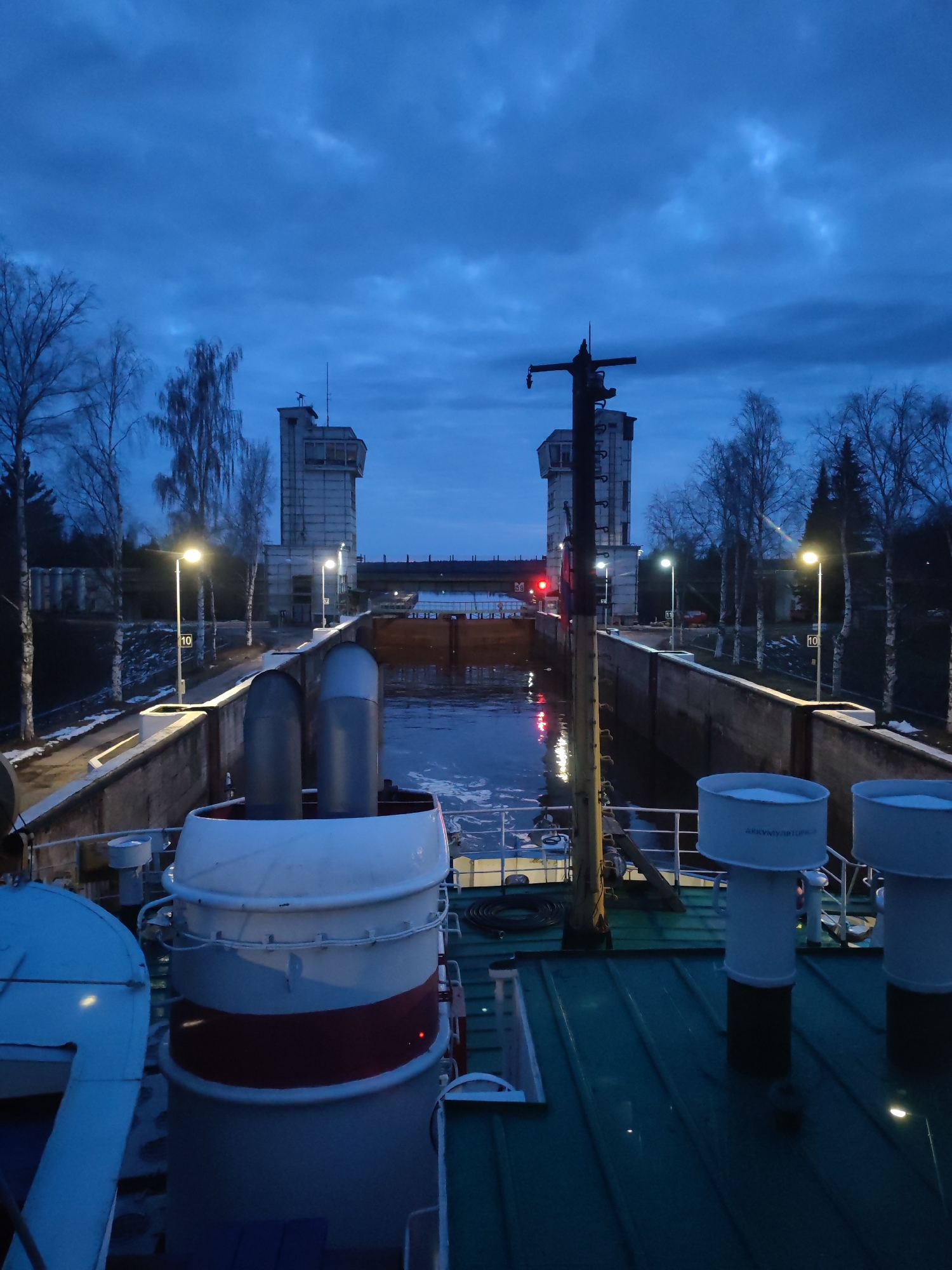 The image depicts a ship within a canal lock at dusk or early night, viewed from the vessel's deck. The sky is a dark, cloudy blue, reflecting the ambient light.
In the foreground, the ship's deck is visible, featuring a large, cylindrical white structure with a red stripe, possibly a funnel or mast base. To its right, several smaller white ventilation stacks or pipes stand on the green deck; one clearly displays the text "AKKUMULYATUR".
The lock itself is defined by concrete walls on both sides, with dark water filling the channel. Bright streetlights illuminate the lock area and a path running alongside the walls. Yellow markers with the number "10" are visible on the lock walls on both sides. A prominent red light is visible near the left side of the lock opening, possibly indicating a signal for the ship.
In the midground and background, two imposing, multi-story concrete control towers flank the far end of the lock, each with visible windows and antennas on top. Bare trees, likely birches, line the banks behind the lock walls, suggesting a cold season. The overall scene conveys a functional, industrial setting for waterway navigation during twilight hours.