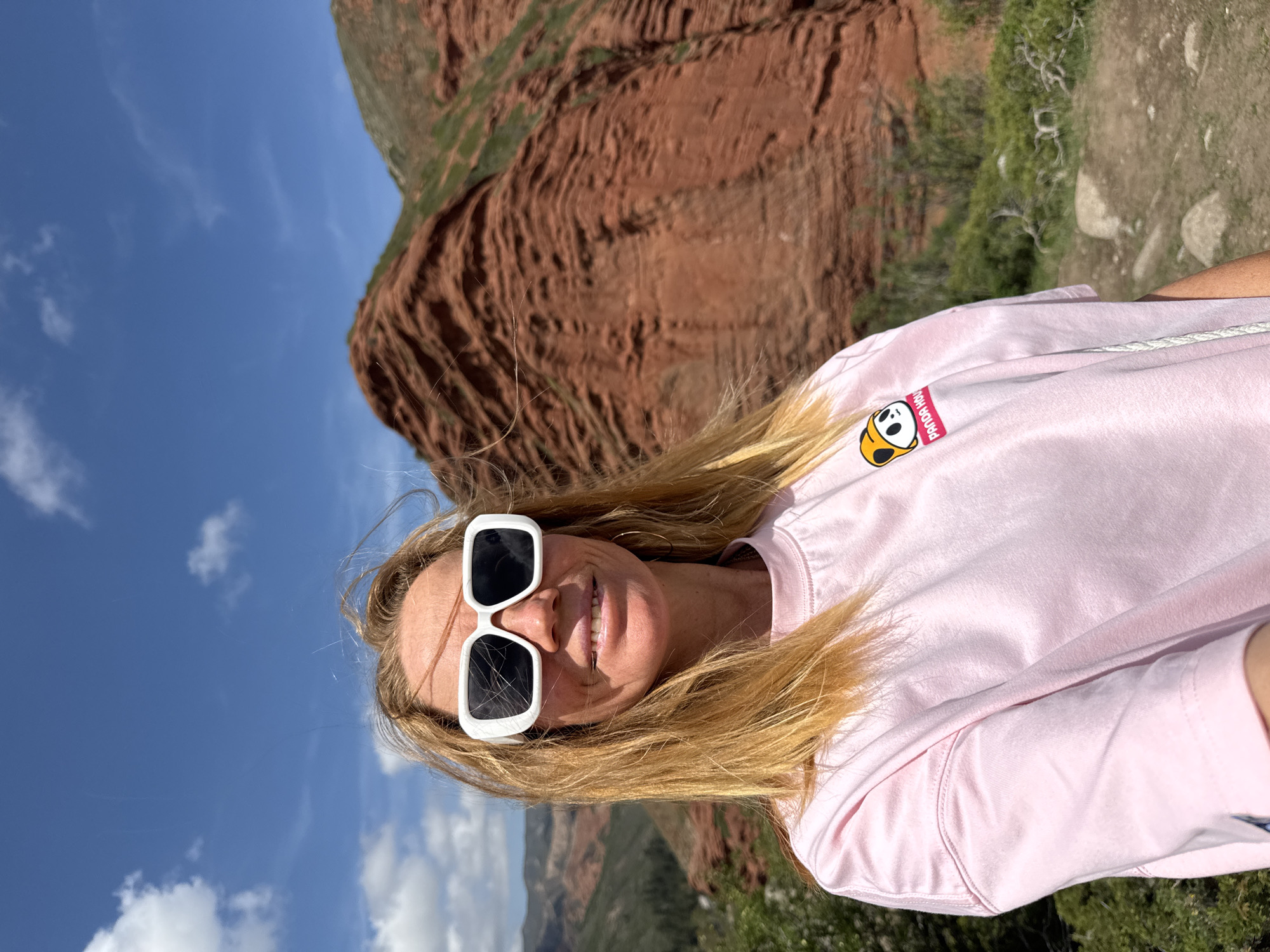 A woman with long blonde hair and white square-framed sunglasses smiles directly at the camera in a vertical, outdoor portrait. She is wearing a light pink t-shirt with a cartoon panda logo on the left chest, which also features the text "Panda HOLA!". The setting appears to be a sunny day, with a clear blue sky and some scattered white clouds visible in the upper and lower portions of the image.
Behind the woman, a dramatic landscape unfolds. Dominating the mid-ground are striking red-brown rock formations, characterized by their layered, almost striated texture, indicative of erosion. These mountains are partially covered with sparse green vegetation in certain areas. To the right of the woman, a dirt path or natural ground is visible, alongside more green bushes and small rocks. The overall impression is that she is standing in a mountainous or canyon-like environment. The image is shot from a slightly low angle, emphasizing the vastness of the sky and the height of the rock formations. Her expression is joyful and relaxed. Given the context, this scene is likely captured in Jeti-Ögüz, Kyrgyzstan.