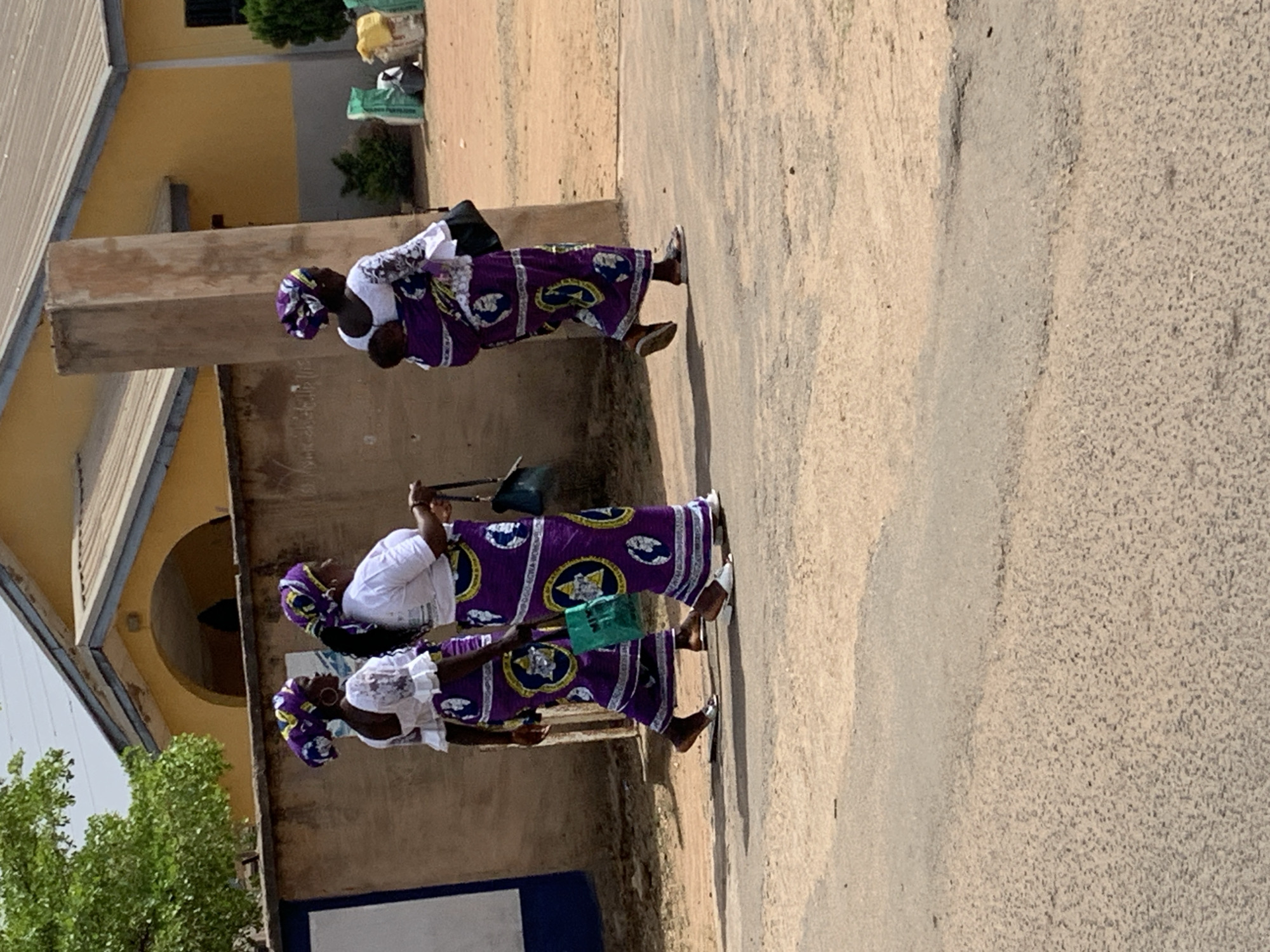 Three women are walking on a paved surface next to a building. They are dressed in traditional African clothing, including white tops, purple skirts with circular designs, and headwraps. The building is two stories tall and has arched doorways and windows. The city of Jos, Nigeria, is the location.