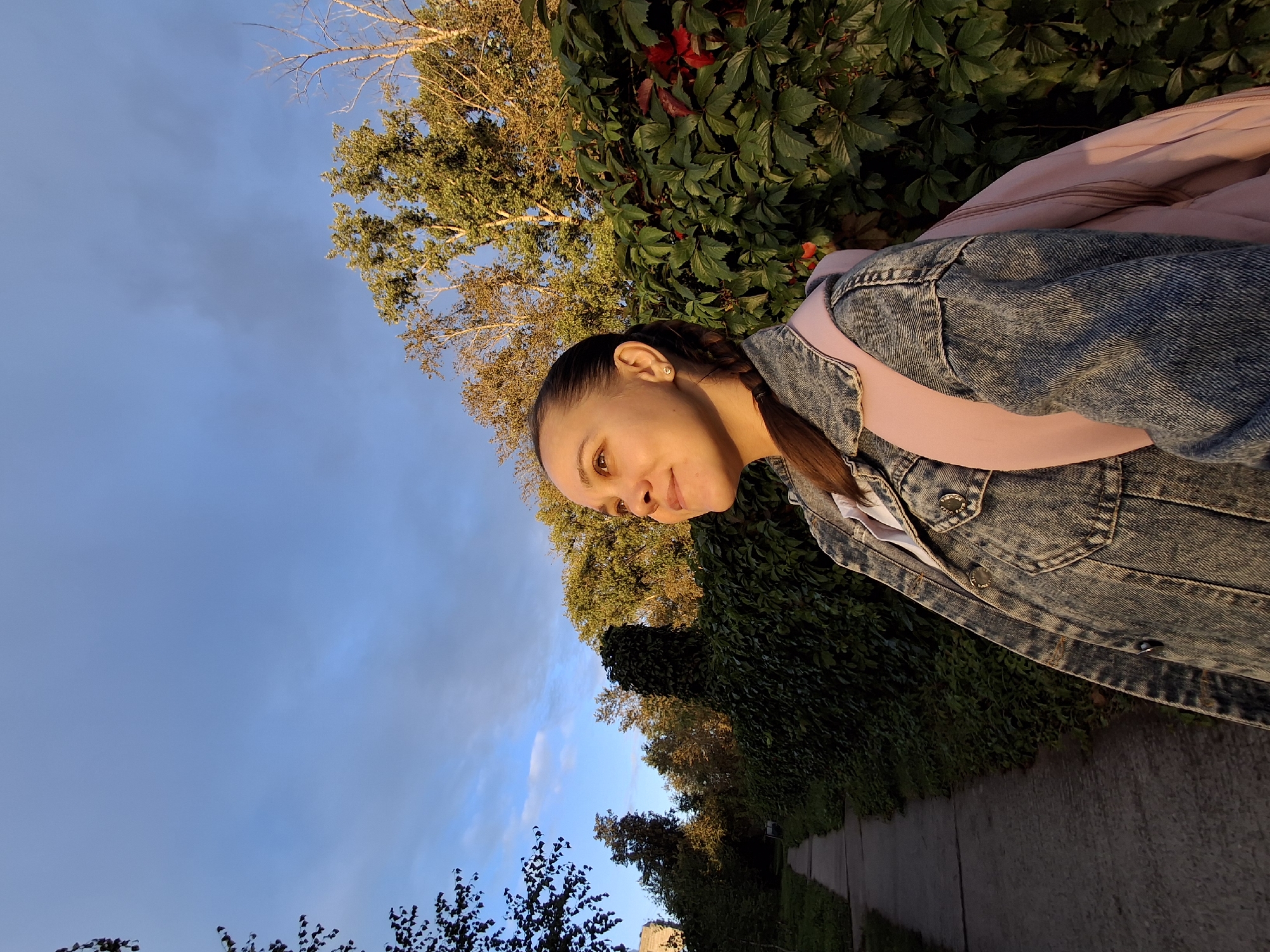 A young woman with a braid is standing on a paved sidewalk next to a dense green hedge and trees. She is wearing a denim jacket over a pink shirt and has a pink backpack on. The sky is blue with some clouds. The sun is setting, casting a warm light on the scene. The location is a street in the city of Chaikovsky, Russia.