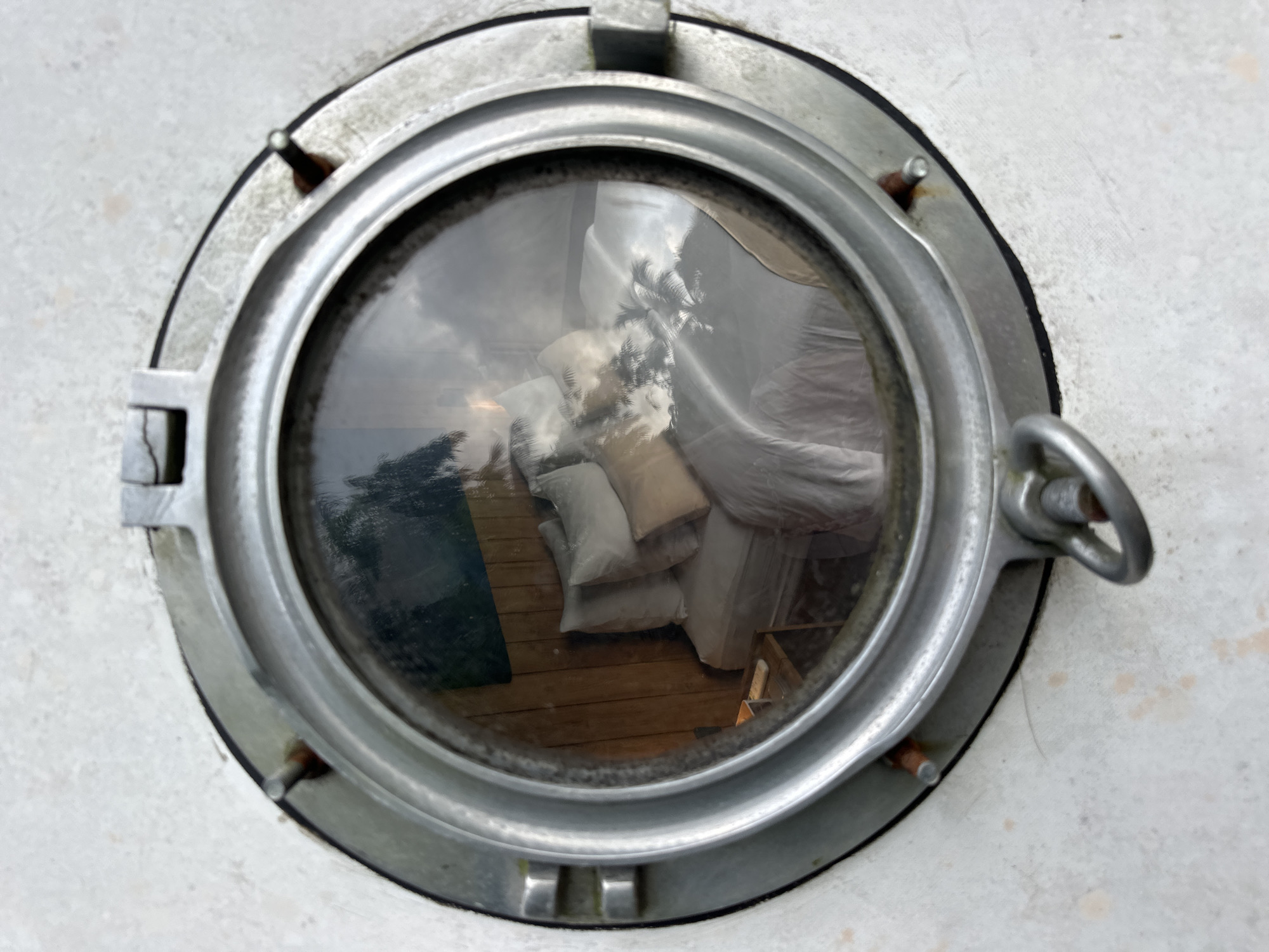 A close-up shot depicts a porthole, likely from a boat or ship, set into a white wall. The circular metal frame of the porthole is weathered, with visible wear and some corrosion. The glass pane is somewhat cloudy, reflecting an interior scene. Within the reflected view, wooden flooring is visible, along with a collection of pillows and what appears to be a mattress or bundled bedding. The reflection also shows a glimpse of foliage, possibly trees or palms, indicating an outdoor setting. The scene suggests an interior space adjacent to an outdoor area, possibly located in Licin, Indonesia, based on the assumption that palm trees are prevalent in that region.