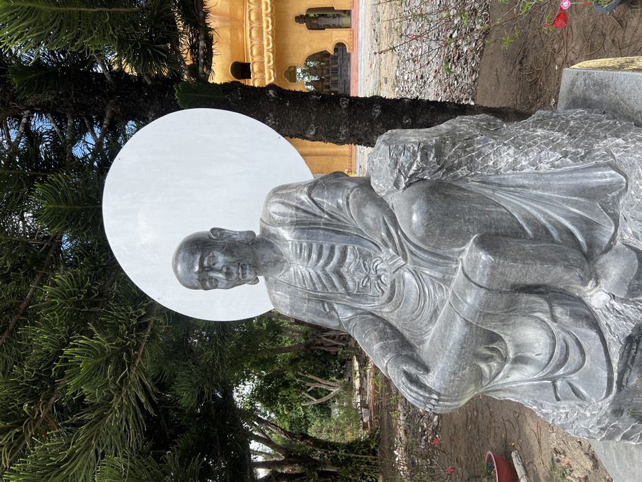 A dark grey stone statue depicting an emaciated, seated figure is centrally positioned outdoors in Đăk Mar, Vietnam. The figure possesses a bald head, elongated earlobes, a visibly open mouth, and a gaunt torso with distinct ribs sculpted beneath draped robes. Its hands rest on its lap, and it is seated on a textured, rock-like base. A large, circular white panel functions as a halo directly behind the figure's head. To the right of the statue stands a large tree with dark, textured bark and dense green foliage. To the left and in the background, additional green trees and vegetation are visible. The ground immediately around the statue consists of bare earth with scattered small stones and sparse red-flowered plants in small pots or bushes. In the background to the right, a multi-tiered building with golden-orange or yellow walls and arched openings is visible, indicative of a temple or pagoda structure. The sky above is partially visible, showing a light blue with some cloud cover.