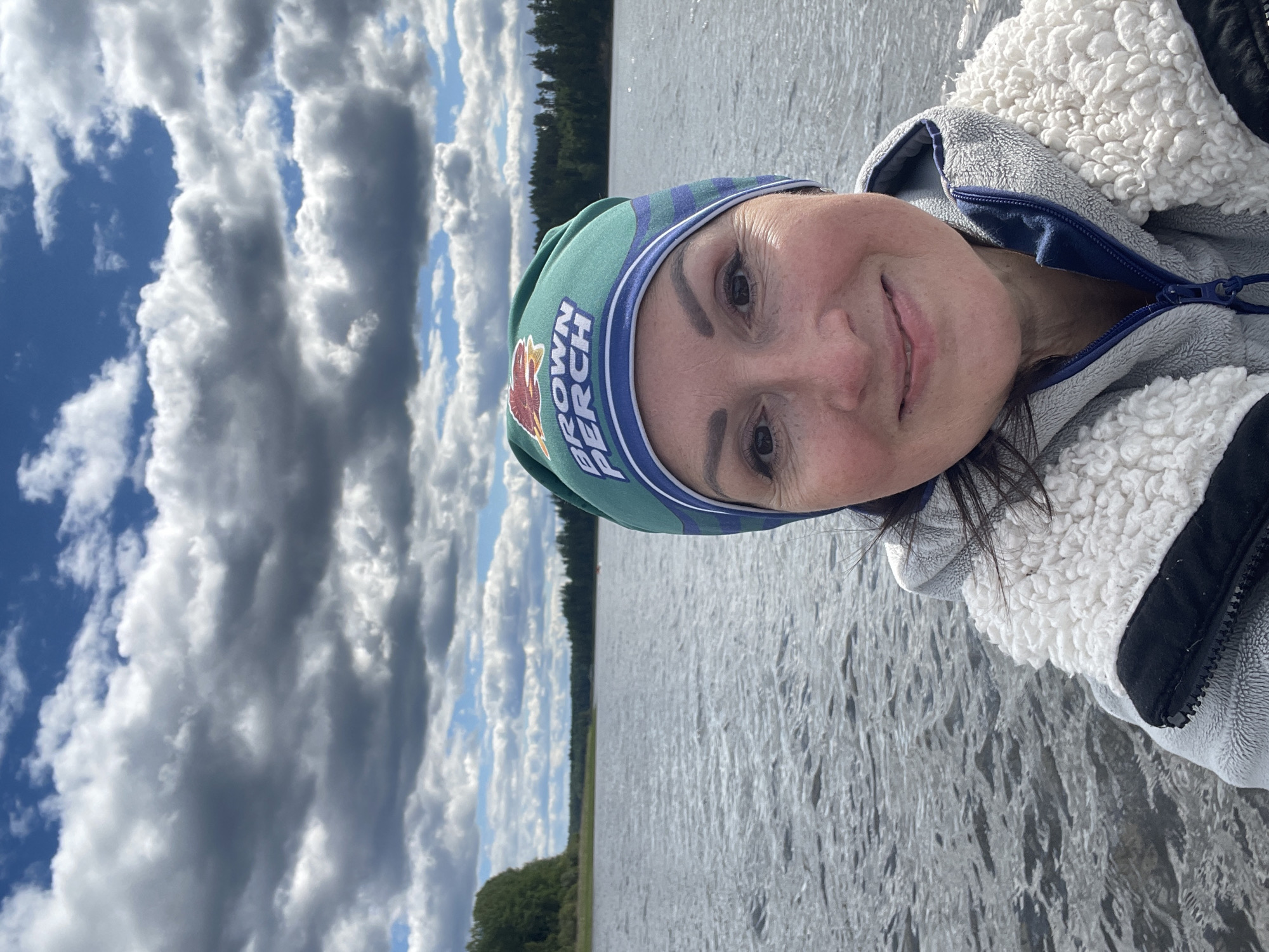 A woman wearing a green beanie with the text "BROWN PERCH" and a fluffy white and gray jacket smiles at the camera in a selfie taken on a body of water. The water has small ripples, and a distant tree line borders the horizon. The sky is filled with dramatic, gray clouds against a blue background.