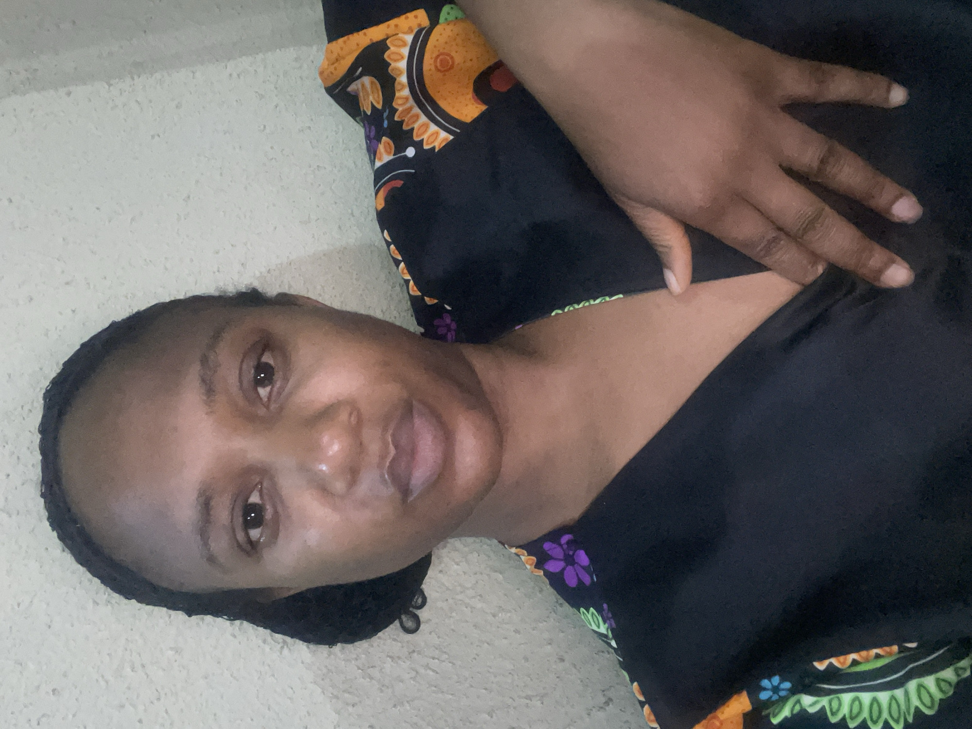 A dark-skinned woman is lying down, looking at the camera. She is wearing a black top with colorful African-inspired patterns around the edges. Her left hand is resting on her chest. The background is a textured, light-colored surface, possibly a wall or floor. There is no information in the image to indicate that it was taken in Jos, Nigeria.