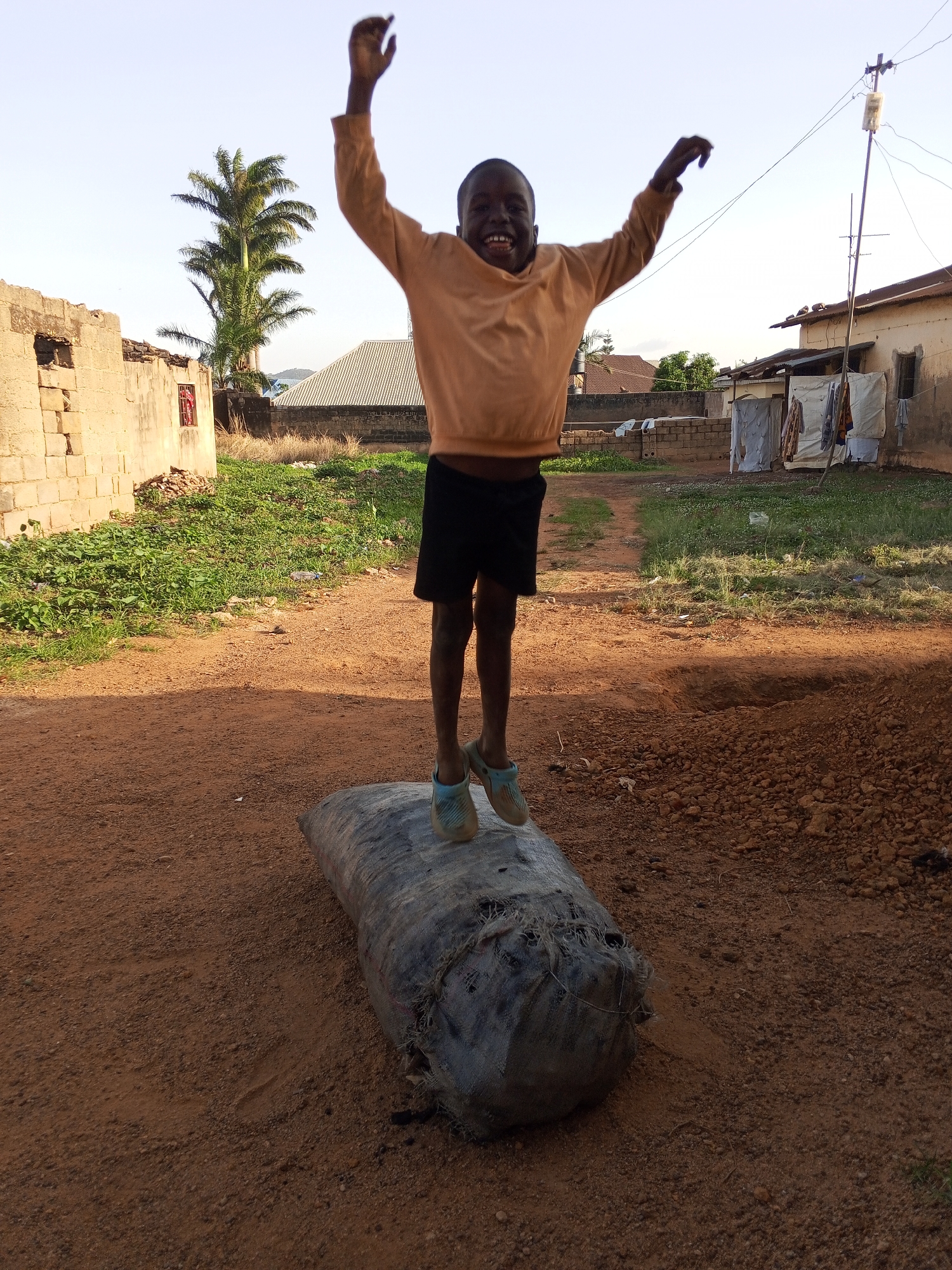 A child is jumping on a large sack in a residential area of Bukuru, Nigeria. The child is wearing a light orange sweater, black shorts, and blue slip-on shoes. The ground is red dirt. In the background are buildings, a palm tree, and clothes hanging on a clothesline. An electrical pole stands near one of the buildings.