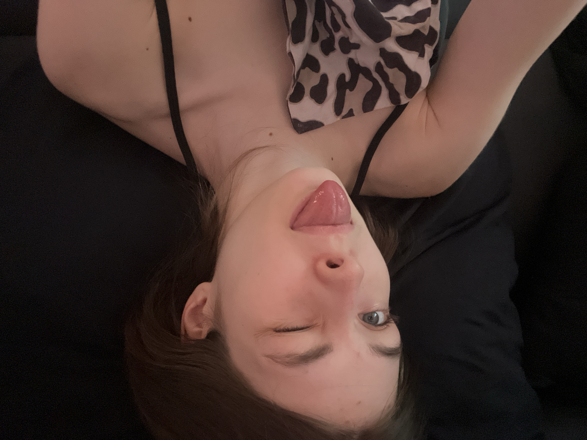 A selfie of a young woman is oriented upside down. She has fair skin, brown hair, and one eye closed in a wink while sticking out her tongue. She is wearing a black tank top. Part of a patterned scarf is visible near her shoulder. The background is dark and indistinct. The location is not discernible from the image.