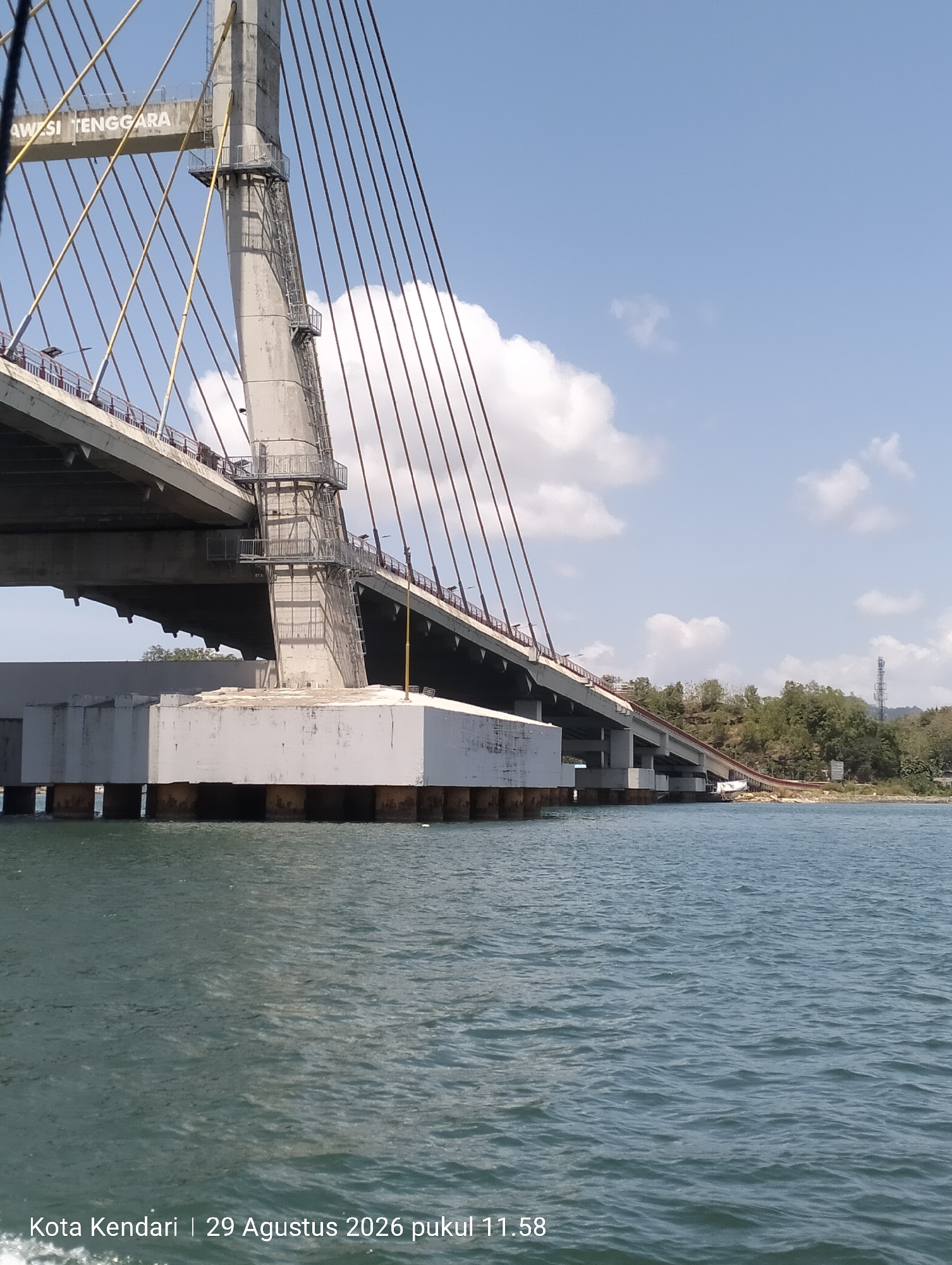 A wide-angle, eye-level, daytime, outdoor photograph captures a cable-stayed bridge over a body of water in Kendari, Indonesia. The bridge, constructed from concrete and steel, features a tall, hexagonal concrete pylon from which numerous cables extend to support the bridge deck. The bridge deck is visible in the mid-ground, with a section of it appearing to be under construction or repair. The supports for the bridge in the foreground are large, rectangular concrete structures resting on submerged pilings. The water in the foreground is a deep blue-green, with gentle ripples on its surface. In the background, a tree-covered hillside rises, with a cell tower visible near the crest. The sky is a clear blue with scattered white clouds. The timestamp overlay indicates the image was taken on August 29, 2026, at 11:58 AM in Kendari. There are no people visible in the image.