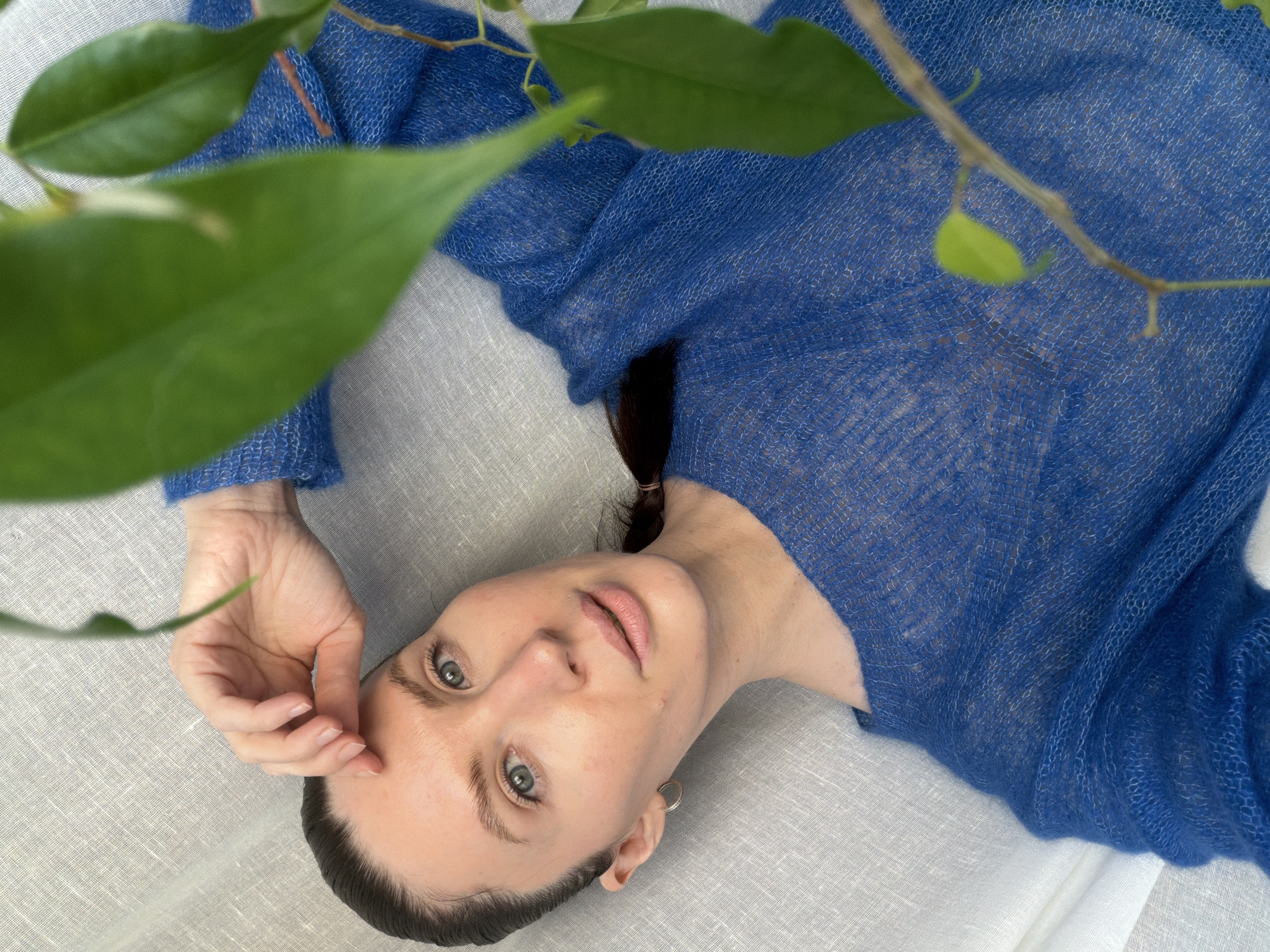 A woman lies on a light grey/white linen surface. She is wearing a blue, loosely woven sweater. Her dark hair is partially pulled back. She looks up at the camera, her right hand resting near her face. Green leaves and a branch are in the upper left of the frame, partially obscuring the scene. The location is unknown, but the request mentioned the city of Perm, Russia. The image composition suggests the photo was taken from directly above the subject.