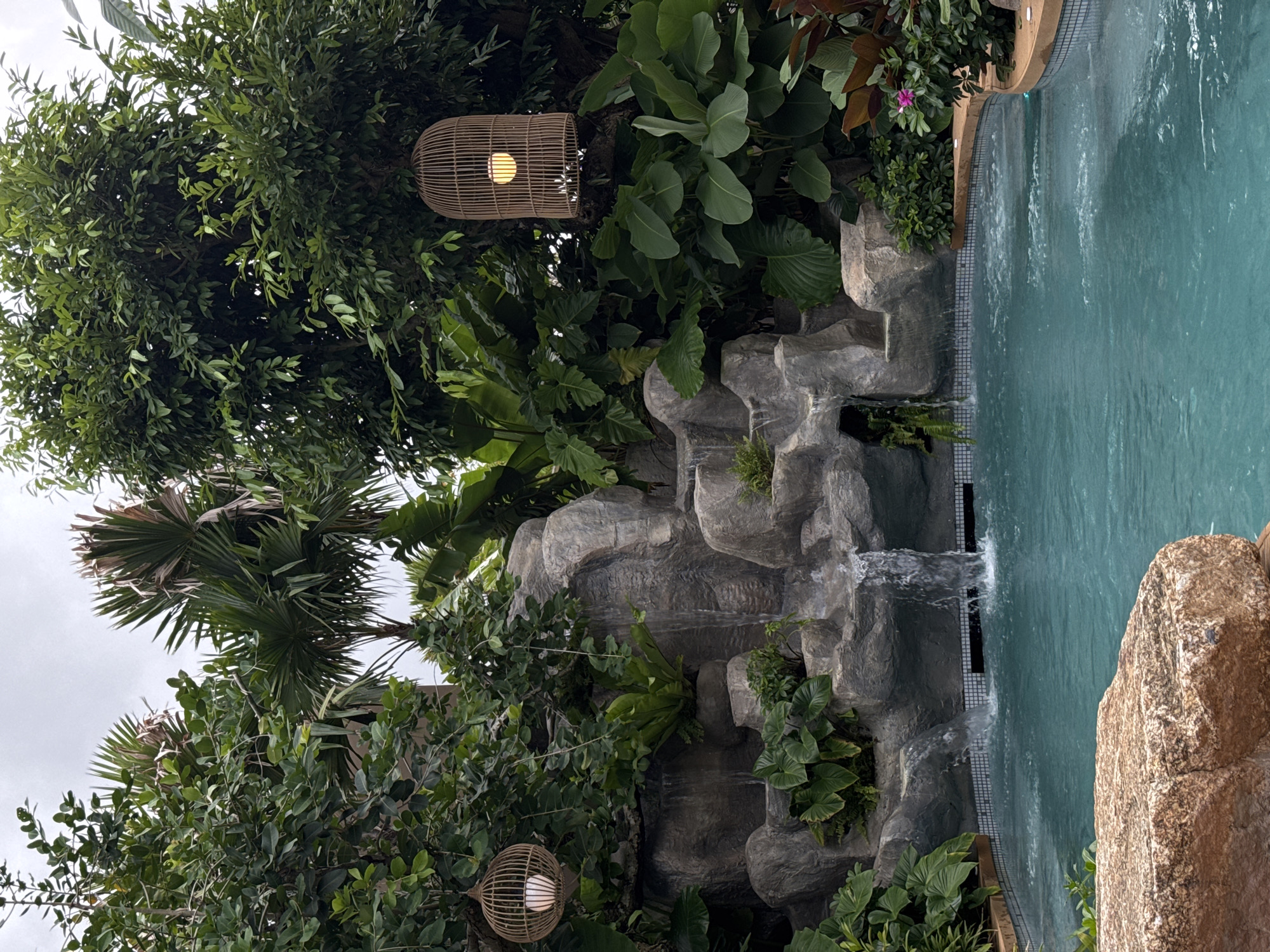 This is a medium shot, eye-level, outdoor photograph of a swimming pool area in Chalong, Thailand. The pool is filled with turquoise water and has a tiled edge. To the left of the pool is a rock formation with a small waterfall cascading into it. Lush green foliage, including palm trees and large-leafed plants, surrounds the rock formation and the pool. Two woven pendant lights hang from the trees, one on the left and one on the right, each with a lit bulb inside. The sky is overcast. The scene is tranquil and appears to be part of a resort or hotel.