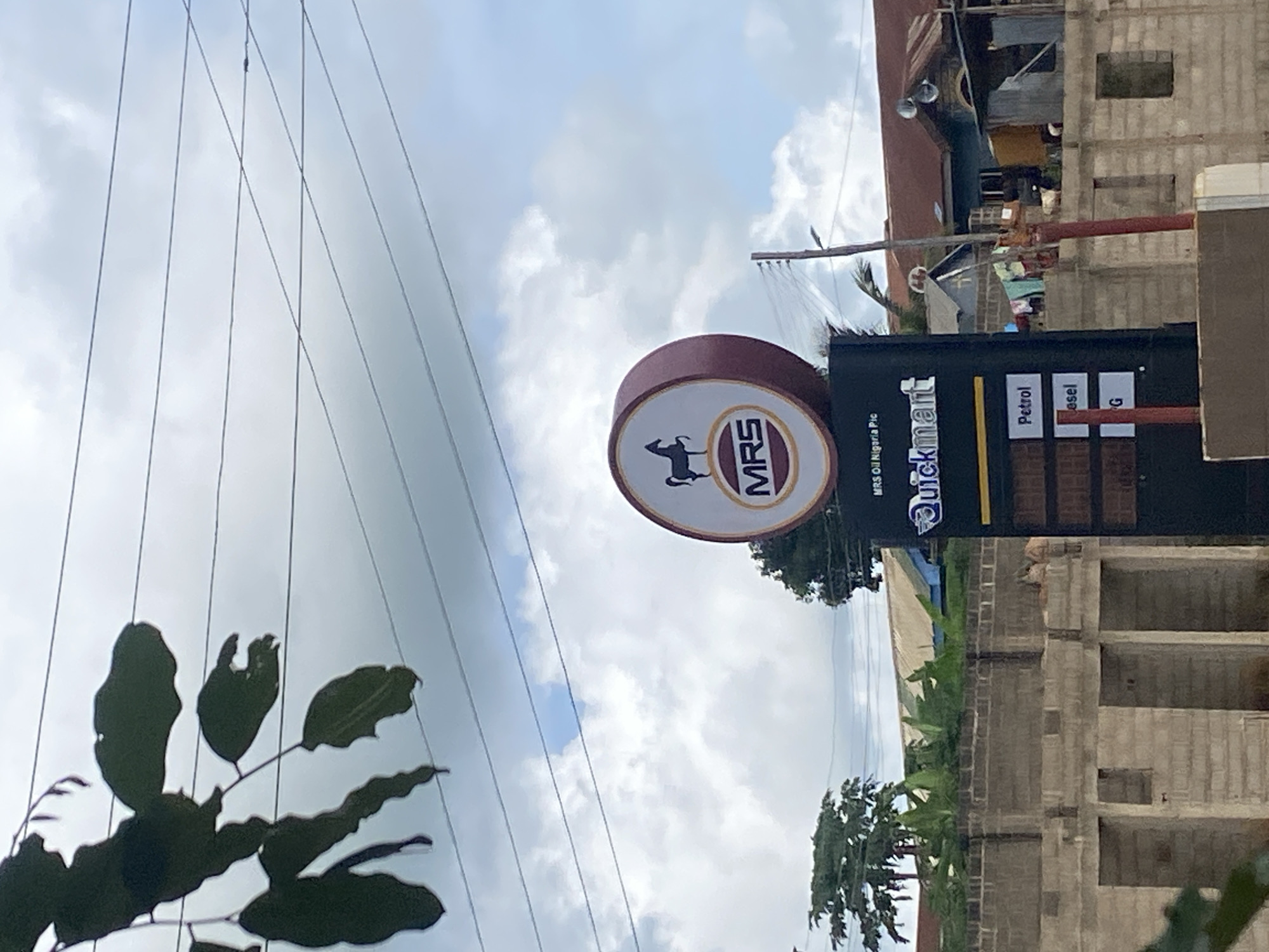 The image, rotated 90 degrees clockwise, captures an outdoor scene featuring a gas station or convenience store sign alongside buildings, power lines, and foliage under an overcast sky.
The central focus is a prominent sign structure. On the left is a circular sign displaying a dark silhouette of an animal, possibly a horse or goat, within a gold-bordered oval. Inside the oval, the letters "MRS" are clearly visible. Below this, smaller text reads "MRS Oil Nigeria Plc." To the right of the circular sign is a rectangular, dark-colored vertical sign with "QuickMart" written horizontally near the top in white and yellow text. Below "QuickMart," there are rectangular panels listing fuel types: "Petrol," "Diesel," and a partially visible label beginning with "G."
In the background, various buildings are visible. Some appear to be concrete structures, possibly residential or commercial, with unfinished elements like exposed blockwork, suggesting construction or renovation. Trees and other green foliage are scattered throughout the mid-ground. Numerous power lines stretch across the upper portion of the frame against a cloudy, bright sky, indicating daytime. Dark-silhouetted leaves are visible in the bottom-left foreground. No people or specific activities are discernible in the image. The scene suggests a roadside or urban environment, consistent with the provided location context of Sasaro, Nigeria.