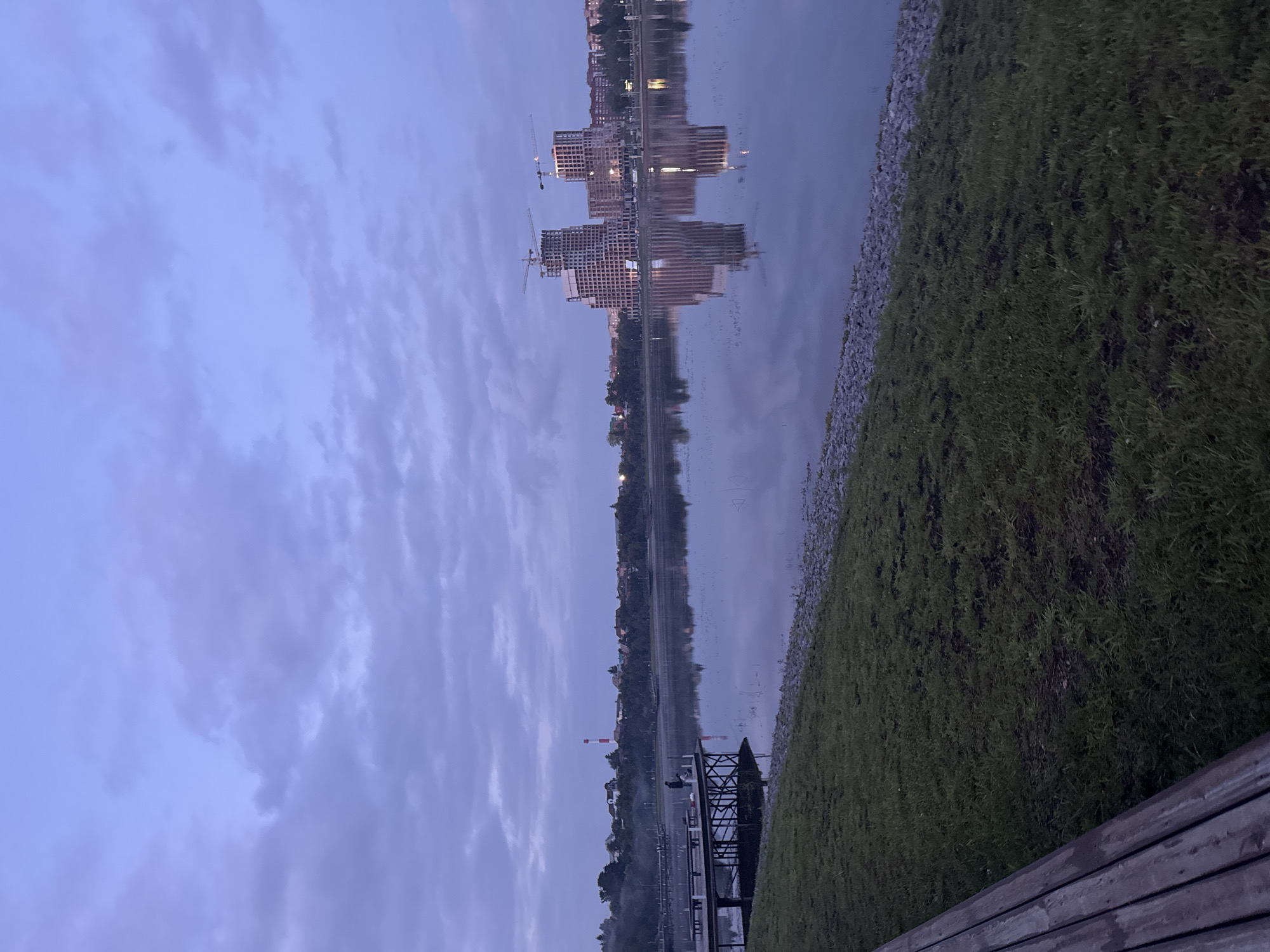 This is a photograph of a lake in Ufa, Russia, taken at dusk. In the foreground, a wooden bench is visible on a grassy embankment. A pier extends into the calm water, where a single person is standing, appearing to fish. On the far side of the lake, a skyline is dotted with trees and buildings. Notably, two multi-story buildings under construction with cranes are prominent in the center of the opposing shore, casting reflections on the water's surface. The sky is a deep blue with scattered purple clouds, and the overall lighting suggests it is twilight.