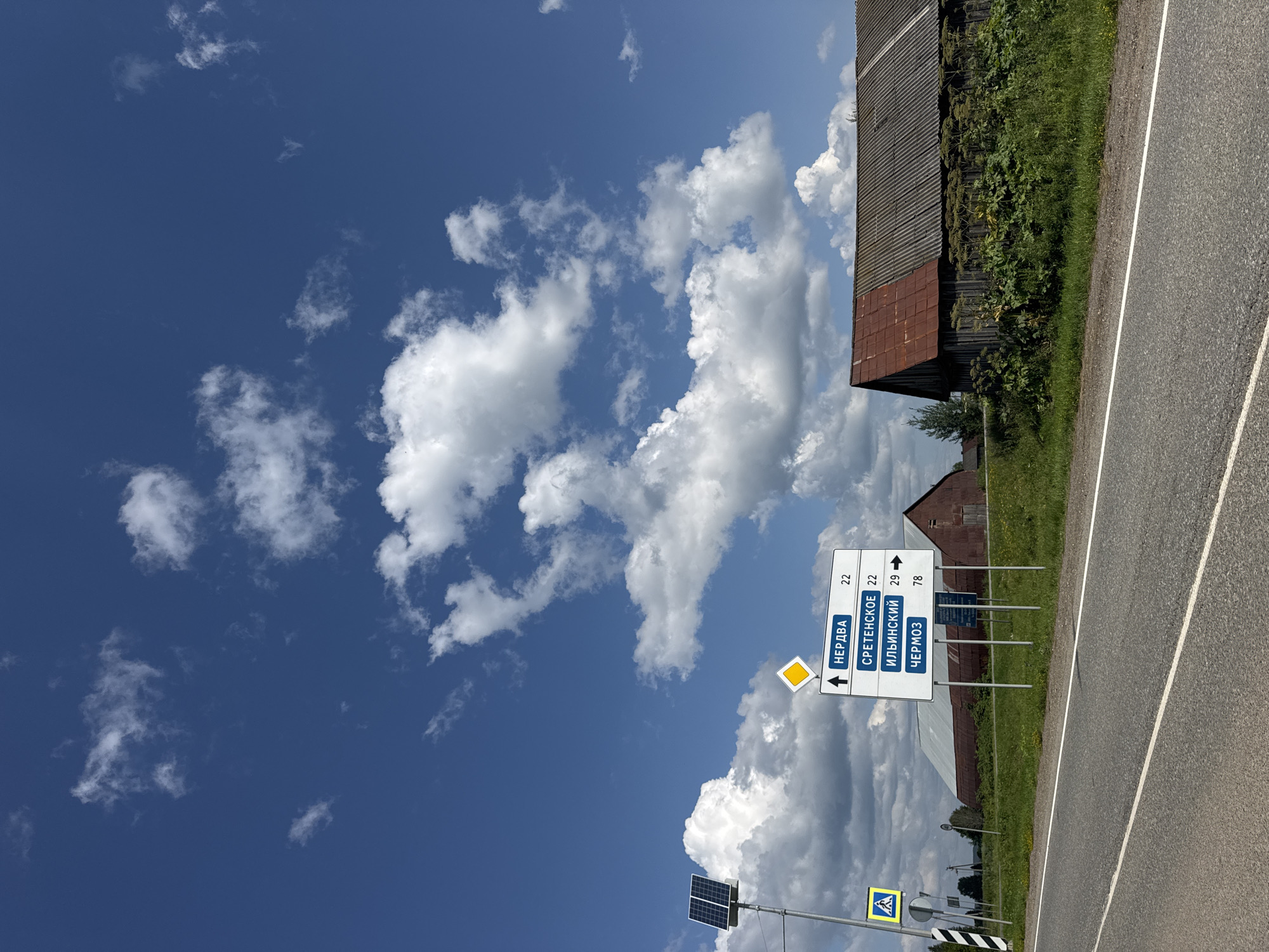 The image captures a rural roadside scene under a bright blue sky with scattered white clouds. A paved road runs diagonally across the foreground, with a white lane divider. On the left side of the road, a signpost displays destinations in Russian: "НЕРДВА 22", "СРЕТЕНСКОЕ 22", "ИЛЬИНСКИЙ 29", and "ЧЕРМОЗ 78". A yellow diamond-shaped sign is mounted above these destination signs, and a pedestrian crossing sign is visible to the left of the destination signpost. A solar panel is mounted on a pole near the signs.
In the background, several old buildings are visible. One building on the right has a dark wooden facade and a rusty corrugated metal roof. Another building behind it is a large barn with a corrugated metal roof and walls. Green grass and vegetation cover the ground around the buildings and along the roadside. The environment appears to be a rural area, possibly in or near the city of Рождественск, Russia, given the road signs. No people are present in the image.