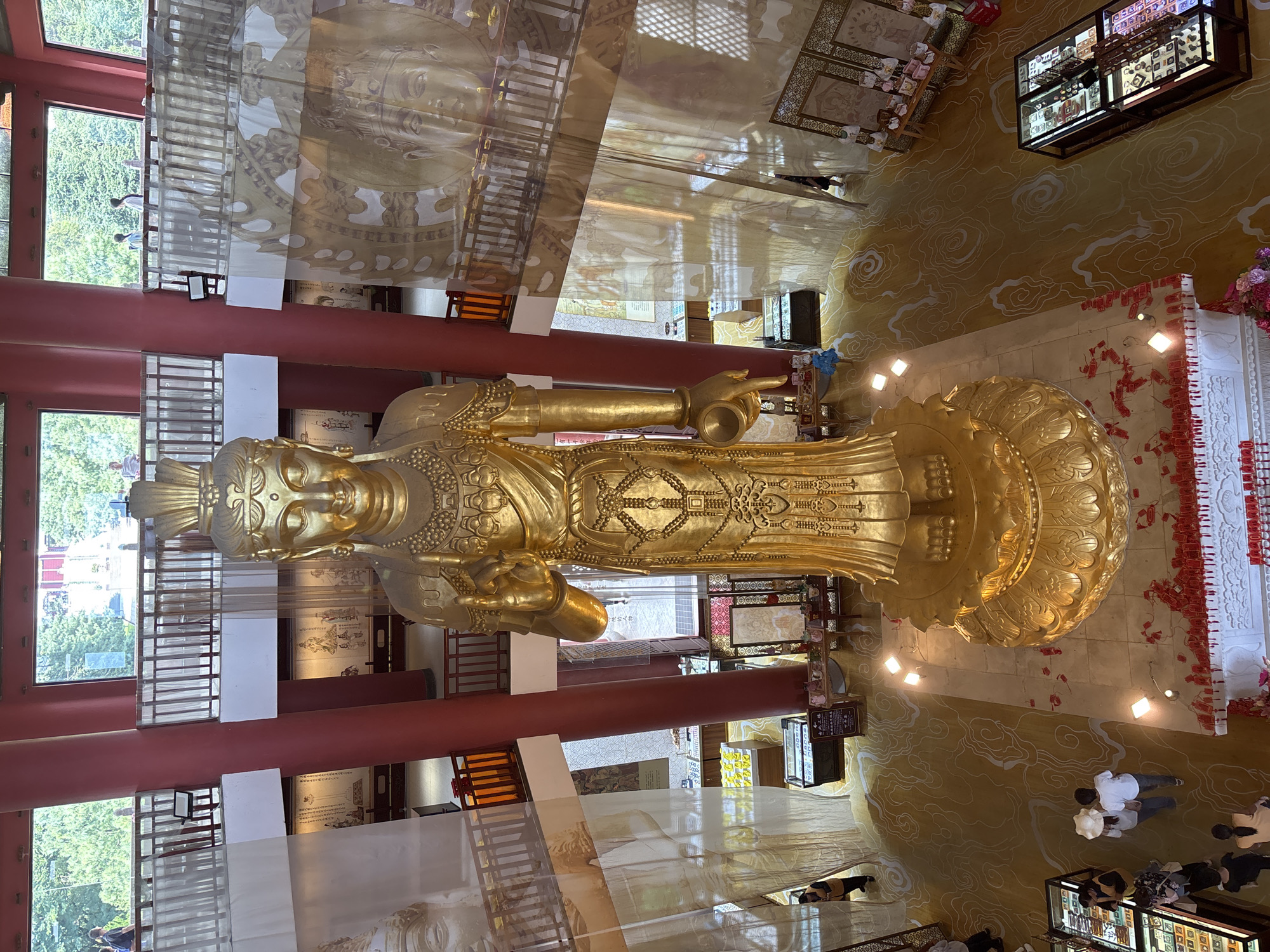 This is an interior shot of a grand hall featuring a towering golden statue of a Buddhist deity, likely Guan Yin, centrally positioned on a lotus pedestal. The statue is adorned with intricate details and jewelry. The hall has high ceilings with red cylindrical pillars supporting balconies on multiple levels. Large windows overlook lush green trees and foliage. Several large, translucent banners depicting Buddhist imagery hang from the upper levels. The floor is a golden carpet with a swirling cloud pattern.
On the ground floor, there are display cases with merchandise, small tables with offerings, and several individuals walking around. A group of two people, a man and a woman wearing hats, are walking away from the camera towards the left. To the left of the statue, a woman in a yellow vest is walking towards the camera.
The overall impression is of a significant religious or cultural landmark in a serene and ornate setting. This location is identified as the Golden Temple (Jindian) in Dali City, China, a famous Taoist temple known for its bronze architecture and scenic surroundings.