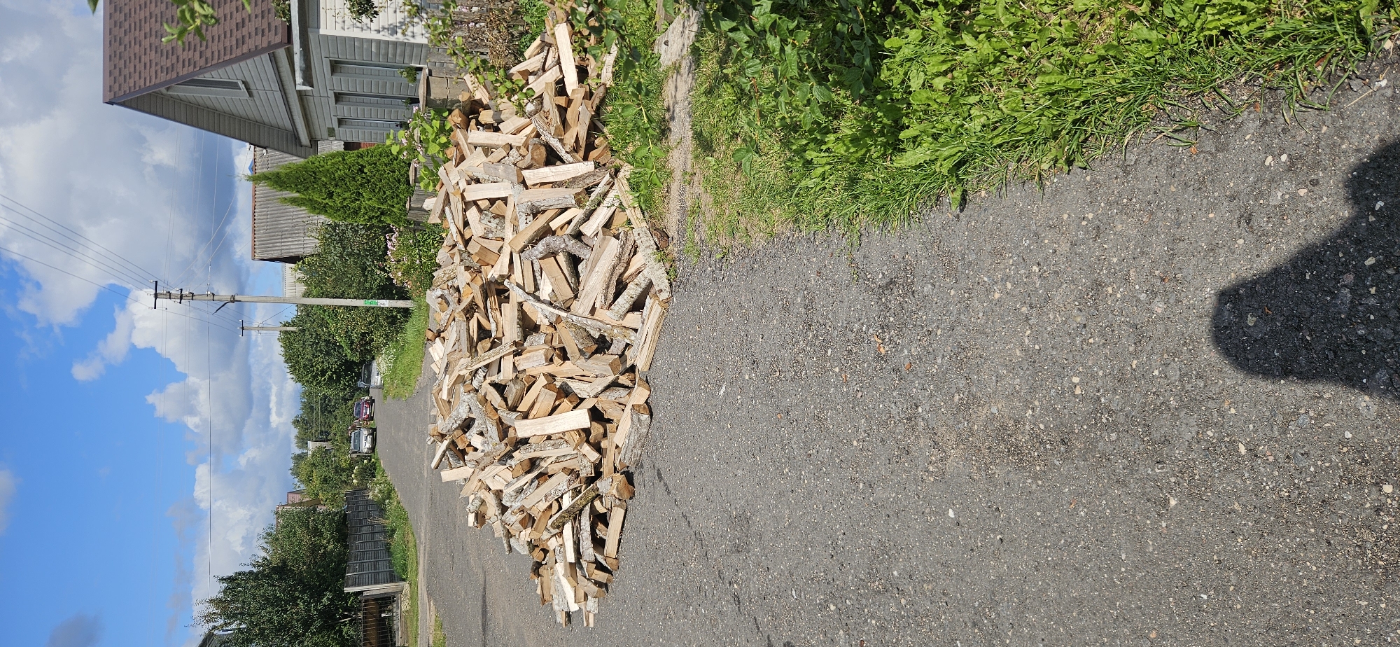This is a full shot, outdoor, daytime image of a pile of chopped firewood on an asphalt road in what appears to be a residential area. The firewood is light brown and stacked in a large, irregular pile in the center of the road. The road is surrounded by green grass on the right side and a gray wooden fence on the left. In the background, there are houses with gray roofs and light-colored siding, trees, and utility poles with wires. Two cars are visible in the distance on the road, and a shadow of a person is cast on the asphalt in the foreground. The sky is blue with white clouds. The image appears to have been taken in the city of Vitsebsk, Belarus, given the architectural style of the houses and the general rural-urban fringe landscape.