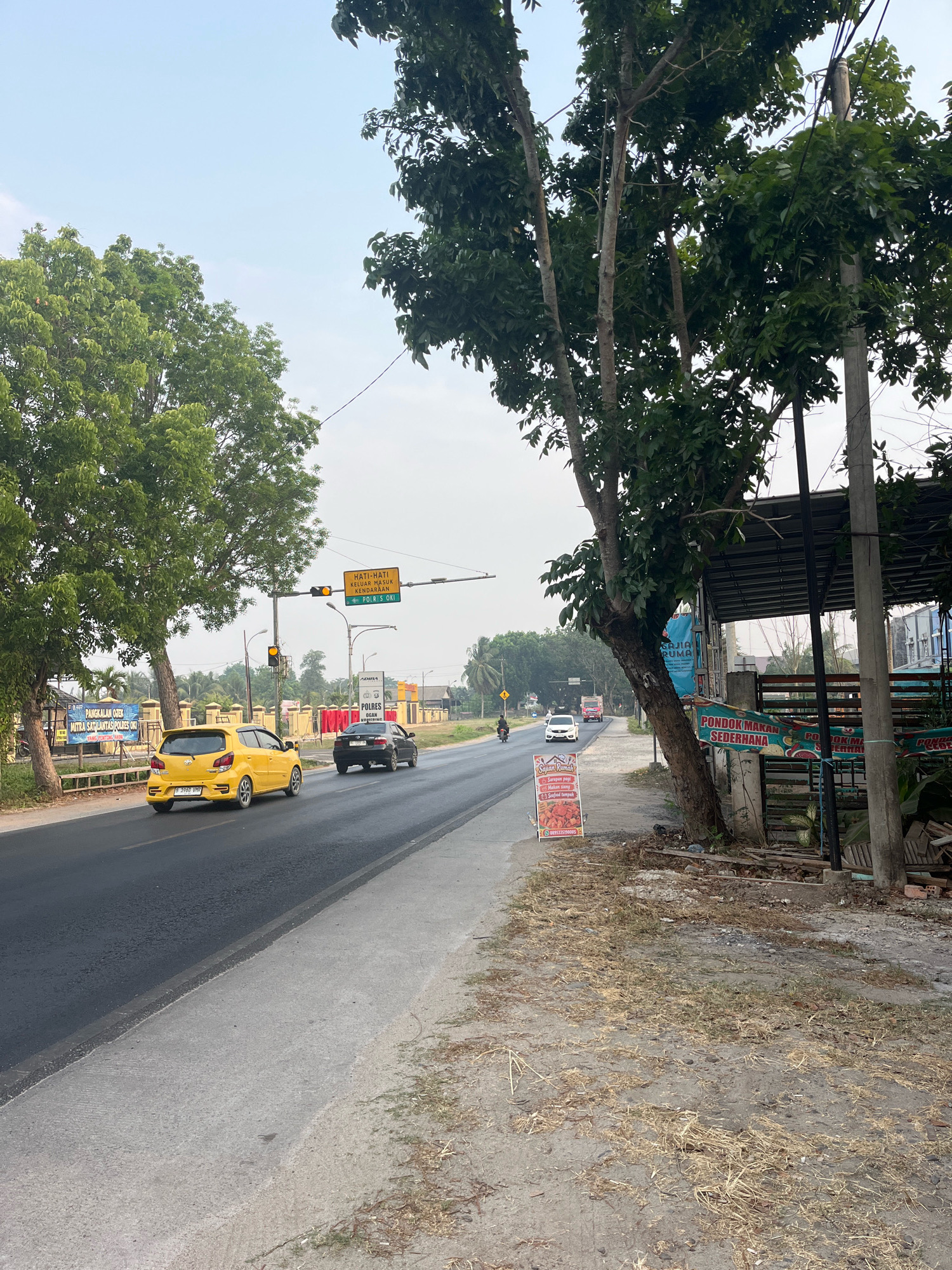 This is a photograph taken from a roadside in Kayu Agung, Indonesia, likely on a street in or near the city. The image depicts a road with two lanes of traffic, with a yellow car and a black car driving on the right side of the road. A traffic light is visible, indicating yellow. On the left side of the road, there are trees and a sidewalk. A sign with Indonesian text and a red border is placed on the sidewalk. In the background, there are buildings and more trees. The sky is overcast. The overall scene suggests a typical day in a town or city, with traffic and commercial activity.