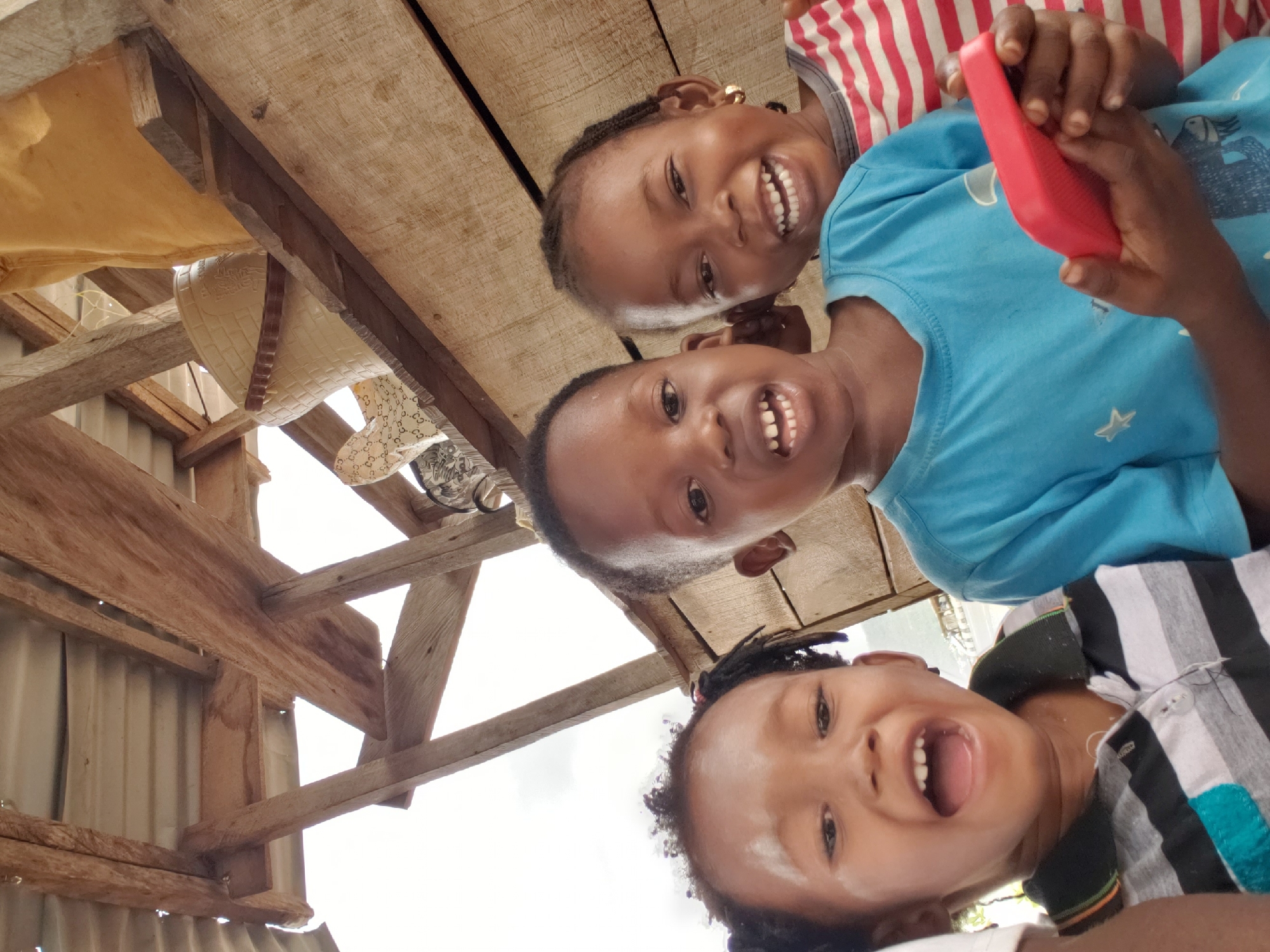 The image features three young children, with dark skin and hair, happily smiling and looking directly at the camera, likely participating in a selfie or a photo taken from a low angle. The child at the bottom of the frame is smiling widely with an open mouth, showing white teeth, and wearing a striped shirt in black, white, and a light blue or teal color.
In the middle of the frame, another child with a wide, toothy smile is wearing a bright blue t-shirt with a faint white design, possibly a star. This child is holding a vivid red smartphone in both hands, suggesting interaction with the device. Above them and to the right, the third child, with braided hair and a gold-colored earring, also smiles broadly, revealing teeth. This child is dressed in a red and white striped top.
The background reveals a rustic overhead structure constructed from wooden planks and beams. Parts of what appears to be a corrugated metal roof or wall are visible on the left side. Hanging from the wooden framework are various items, including a light beige fabric and a patterned white cloth. A beige, textured container, possibly a basket or light fixture, is also suspended from the beams. The bright lighting suggests it is daytime. The children's joyful expressions convey a sense of happiness and playfulness in what appears to be an indoor or covered outdoor setting in Adankolo, Nigeria. No visible text is present.