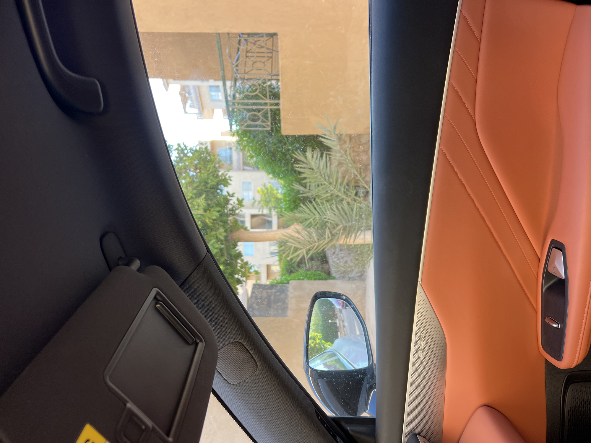 The image shows the interior of a car, looking out the passenger window. The car's interior features orange leather upholstery on the door panel and a black headliner with a sun visor. The window reveals an outdoor scene with a tree, green foliage, and buildings in the background. A side-view mirror on the car reflects a street with other vehicles. The outdoor environment appears to be an urban or suburban area with a warm, sunny climate, consistent with a location in Egypt. The overall impression is a snapshot of a car journey on a bright day.