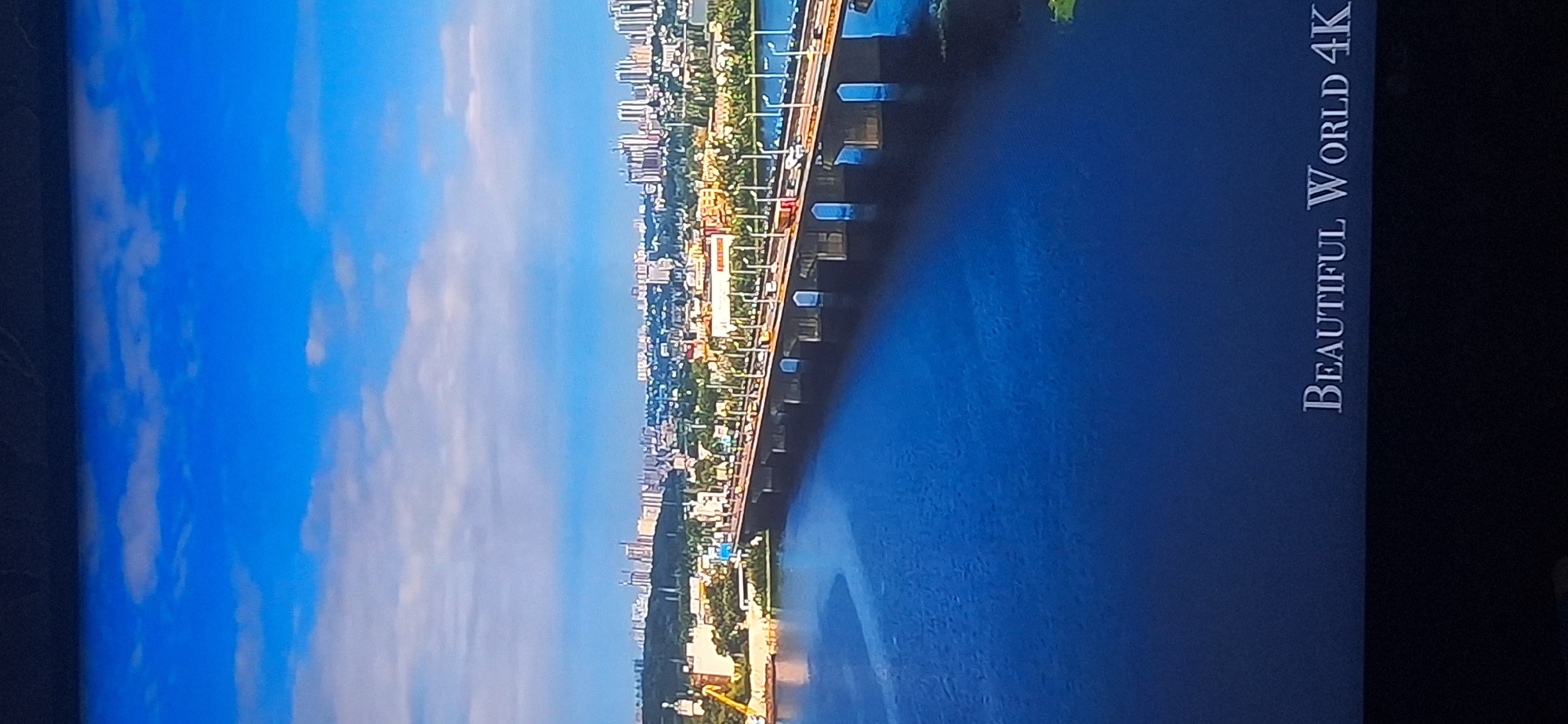 The image is an aerial view of a city, likely Bila Tserkva, Ukraine, dominated by a wide, blue river in the foreground. A long, multi-lane bridge stretches across the river, with vehicles and streetlights visible on its surface. The far bank of the river is lined with a dense urban landscape featuring numerous buildings of varying heights, including many modern high-rise structures and construction cranes, interspersed with greenery. The sky above is a vibrant blue with scattered white clouds. In the bottom right corner, the text "BEAUTIFUL WORLD 4K" is displayed. The overall scene depicts a bustling cityscape integrated with a significant riverine feature.