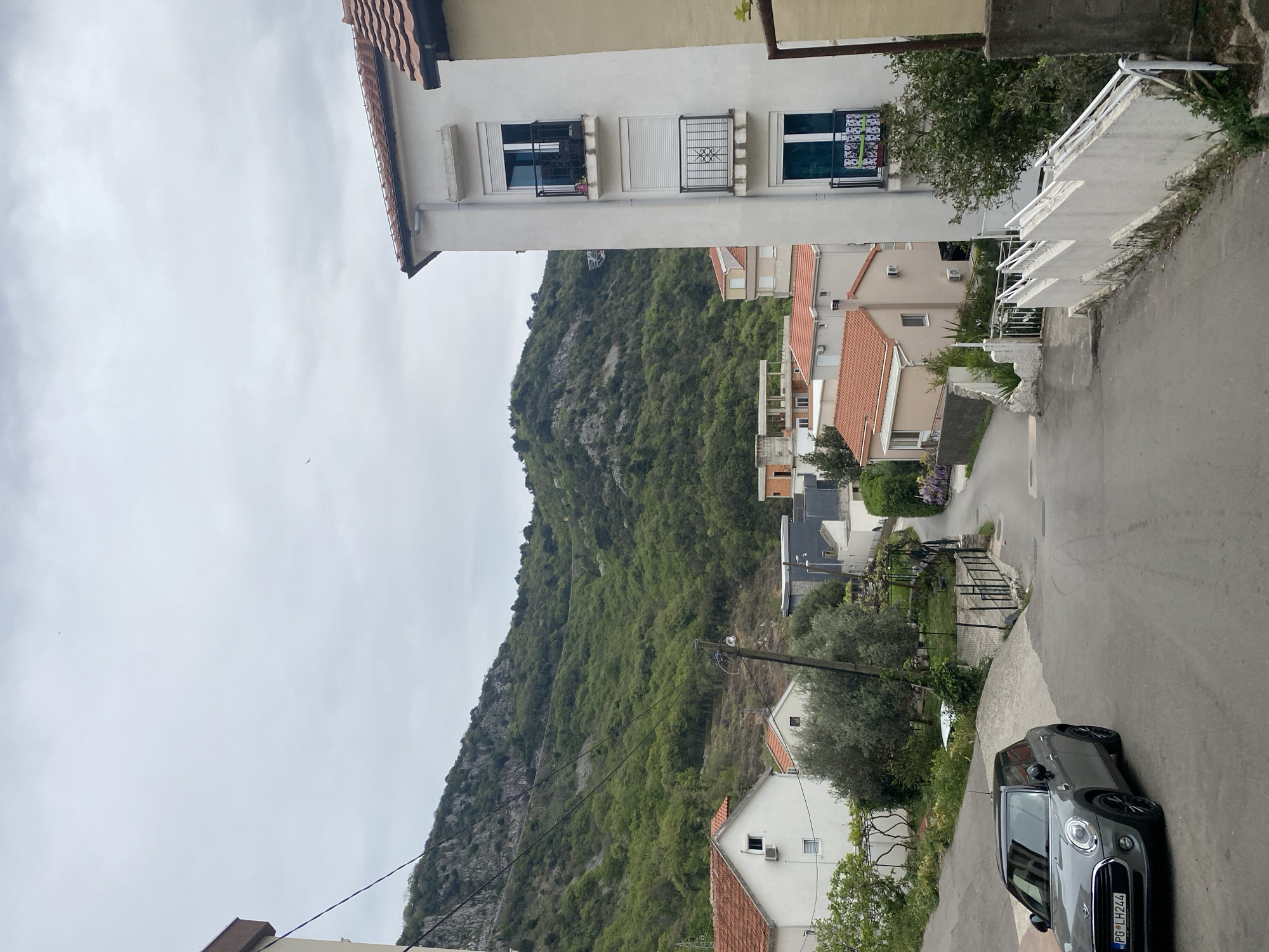 The image shows a street scene in what appears to be a residential area in or near Budva, Montenegro. The view is along a narrow, paved road leading downhill. On the right side of the road, a gray Mini Cooper is parked. Houses with red-tiled roofs and white or light-colored walls line the road on both sides. The architecture is typical of the Mediterranean region. In the background, a large, green, rocky mountain dominates the view. Overhead power lines are visible. The sky is overcast.