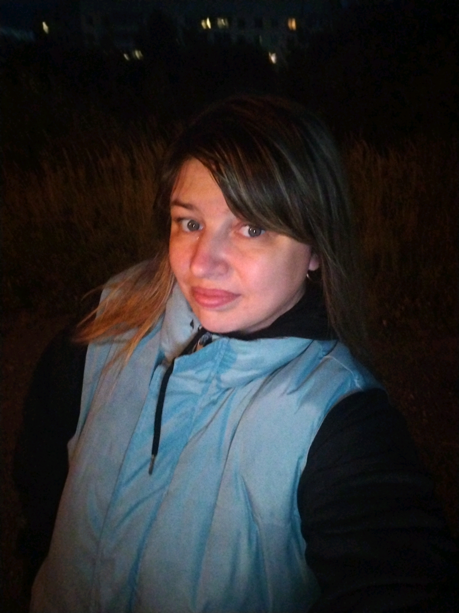The image is a close-up, selfie-style photograph of a person from the chest up, taken outdoors at night. The background is dark and out of focus, with some distant, blurred lights that could indicate a city or artificial lighting. The person appears to be in a natural setting with tall grass or reeds visible behind them.
The subject is a woman with fair skin, light blue eyes, and shoulder-length brown hair with lighter streaks. She is wearing a light blue puffer vest over a black garment, possibly a jacket or sweatshirt, with a black collar visible. The vest has a drawstring closure with a metal pull. Her facial expression is neutral to slightly melancholic.
There are no specific landmarks or indicators in the image that would allow for identification of the location within Yaroslavl, Russia. The general environment suggests an outdoor, possibly somewhat wild or uncultivated area on the outskirts of a populated area, given the distant lights. The time of day is clearly night due to the darkness and artificial lighting.