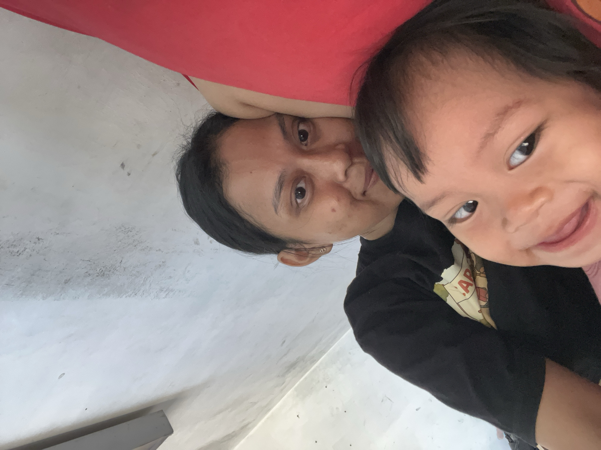 The image depicts a close-up, low-angle selfie of two individuals, seemingly a mother and child, lying down. The child is in the foreground, on the left, with their face angled towards the camera, smiling. Their dark hair frames their face, and their eyes are open and looking forward. The adult is positioned behind and to the right of the child, with their head resting on their arms. Their eyes are open and looking towards the camera. Both individuals are wearing dark-colored clothing, with the adult wearing a black t-shirt, and the child wearing a pink top. They are lying on a red surface, likely a bed or cushion. The background consists of a plain, textured wall that appears to be a light gray or white color, with some minor blemishes and smudges visible. The lighting suggests an indoor setting, possibly a bedroom, with natural light illuminating the scene. The image has been rotated 90 degrees clockwise. The provided context suggests the location is Kecamatan Pati, Indonesia, which is a subdistrict within the Pati Regency in Central Java province.