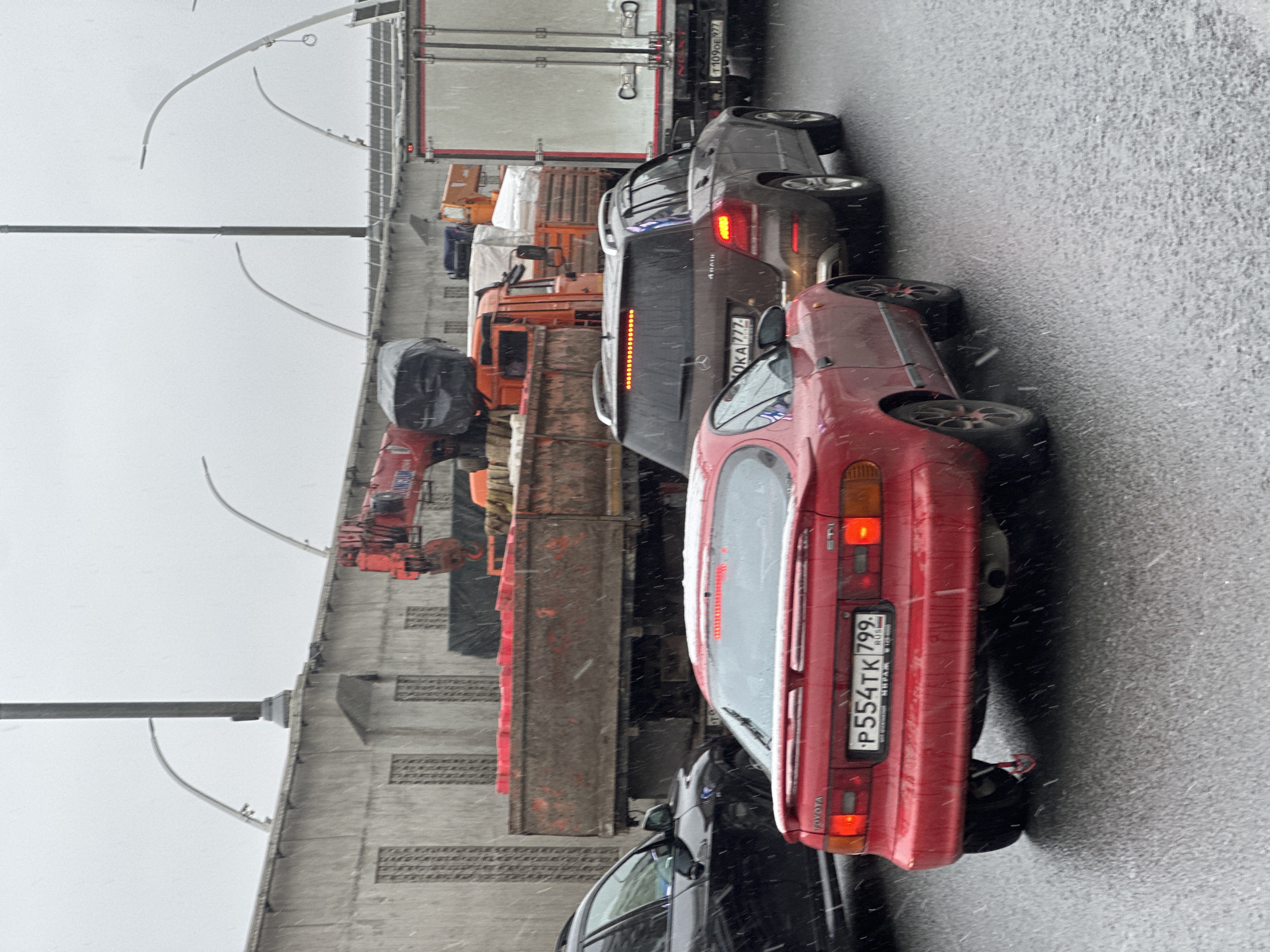 The image depicts a traffic jam on a bridge in Moscow, Russia, during a snowfall. A red Toyota car is closest to the camera, followed by a black car, a dark grey Mercedes-Benz SUV with license plate "OKA 777", a truck loaded with wooden pallets, and other trucks further ahead. The bridge's infrastructure, including support columns and arched streetlights, are visible on the left side of the image. The sky is overcast.