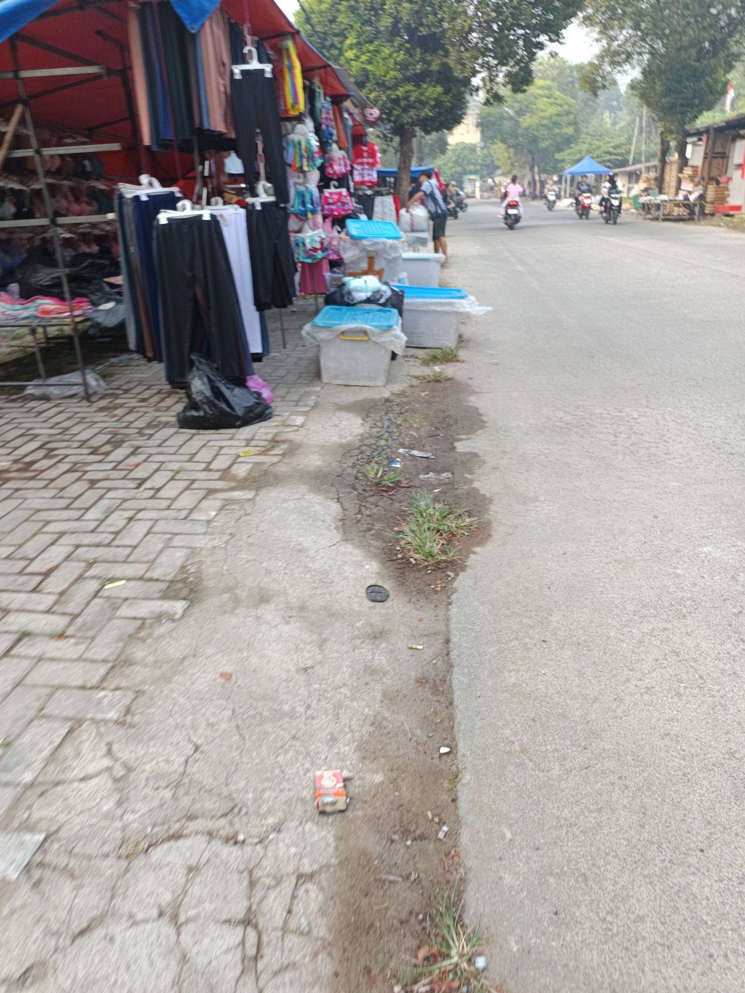 The image depicts a street scene in Sodong, Indonesia, likely on a market day. On the left, a stall displays various clothing items, including pants and shirts, hung from racks. Several large, light blue plastic containers are placed on the ground near the stall, some with lids. A black plastic bag lies on the pavement in front of the stall. The pavement is made of interlocking bricks. The street itself is paved with asphalt and shows signs of wear and tear, with some water pooling and debris visible. Several people are seen on motorcycles further down the street, with more stalls and trees lining the roadside. The environment appears to be a bustling outdoor market.