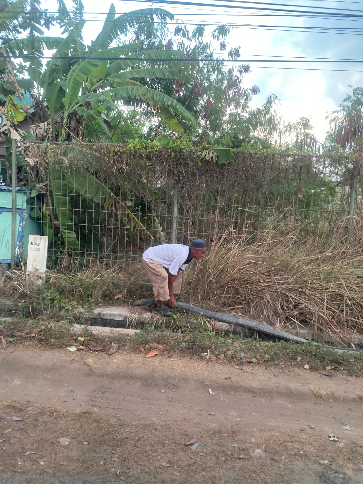 A medium-shot, eye-level, outdoor, full-body photograph shows a man in Banjar Sari, Indonesia, bending over a black pipe near a chain-link fence covered in dry vines and some green plants. The man is wearing a white short-sleeved shirt, tan pants, a blue head covering, and dark sandals. He is positioned on the right side of the frame and is facing the pipe, appearing to be working on it. Behind the fence are large banana tree leaves and other greenery. To the left of the man, a white post with "RMJ 2023" inscribed on it is visible. The ground in front of the man is a dirt path with some dry grass and debris. A concrete gutter runs along the base of the fence. Power lines are visible overhead against a cloudy sky.
