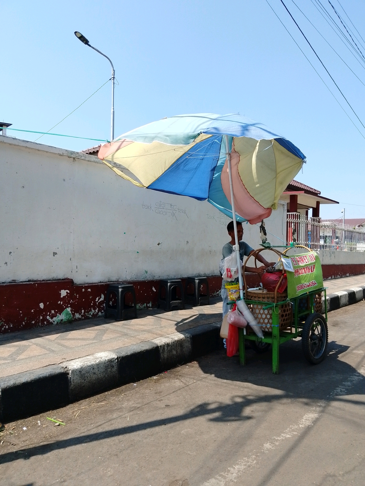 A street vendor stands beside his cart, which is covered by a colorful umbrella. The cart is laden with goods, including what appear to be drinks in plastic cups and bags. A green sign on the cart displays text and a phone number. The vendor, a man wearing a light-colored shirt, is partially obscured by the cart and is engaged with its contents. He is positioned on a paved sidewalk adjacent to a street. Behind him, a white wall with some graffiti and peeling paint is visible. Several black plastic stools are placed against the wall. In the background, a building with a white fence and a red-tiled roof can be seen. The scene takes place outdoors under a clear blue sky, suggesting it is daytime. The overall impression is a typical street scene in a city, possibly a marketplace or a busy thoroughfare. Based on the presence of the sign with Indonesian text "ES BASKET ANYA MAKMUR" and a local phone number, the location is likely Pasuruan, Indonesia.