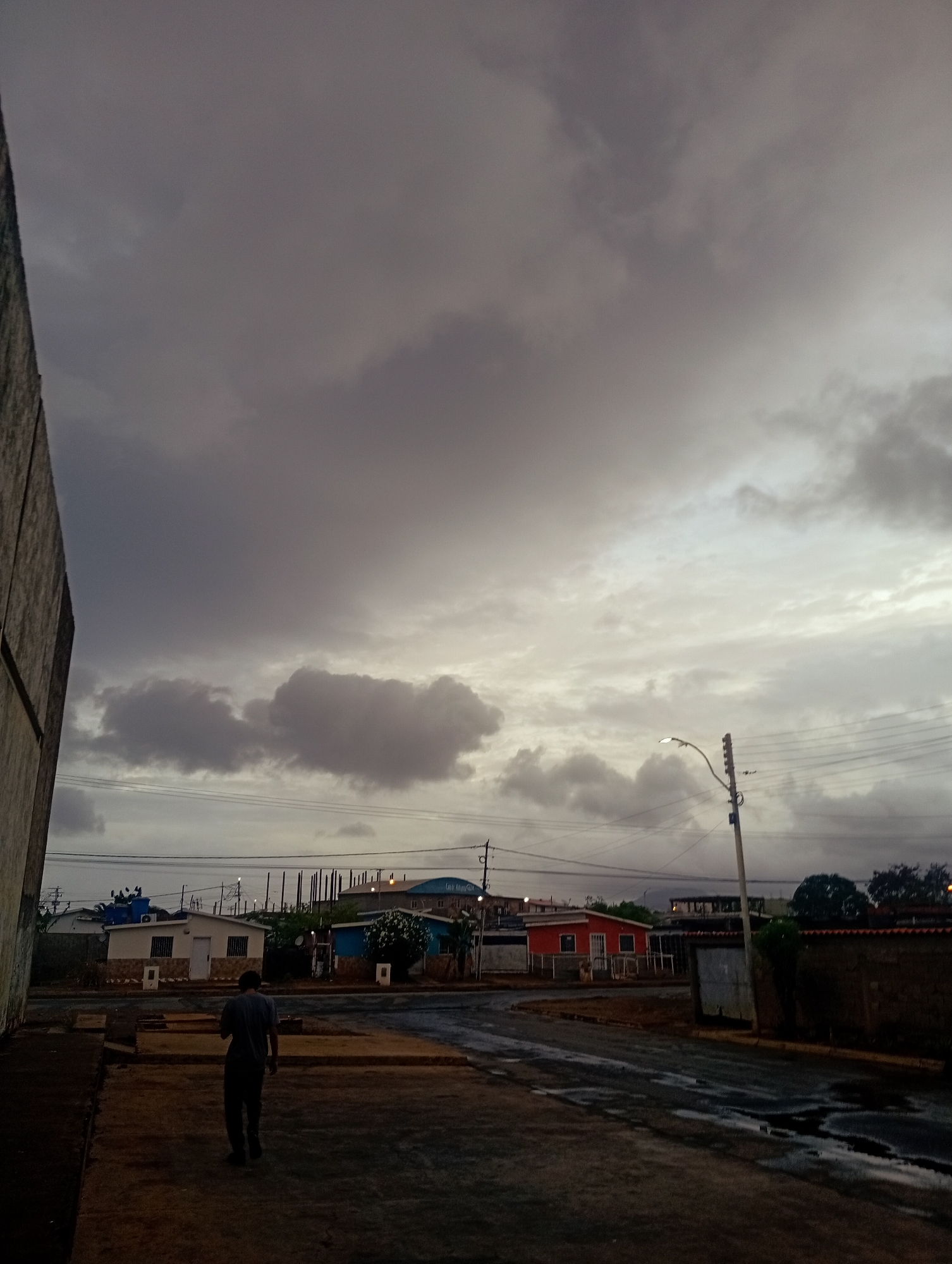 In Juan Griego, Venezuela, a person walks away from the camera on a dark, wet road, likely after rain. The sky is overcast with heavy, gray clouds. Several small, colorful houses, painted in white, blue, and red, are visible in the background, along with utility poles and electrical wires. A street lamp is illuminated. The scene suggests an evening or pre-dawn setting, given the dim lighting and cloudy conditions.