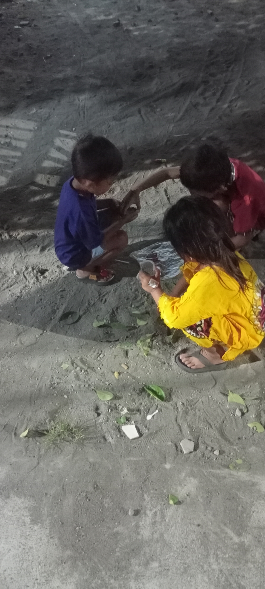 In Gempolklutuk, Indonesia, three children are gathered on a dirt ground. One child, wearing a blue shirt and sandals, is crouching and examining something on the ground. Another child, wearing a red shirt, is leaning over and also looking at the ground. The third child, in a yellow shirt with a colorful pattern, is also crouching and holding a small object. Several green leaves and fragments of white material are scattered on the ground around them. The lighting suggests it is either evening or night.