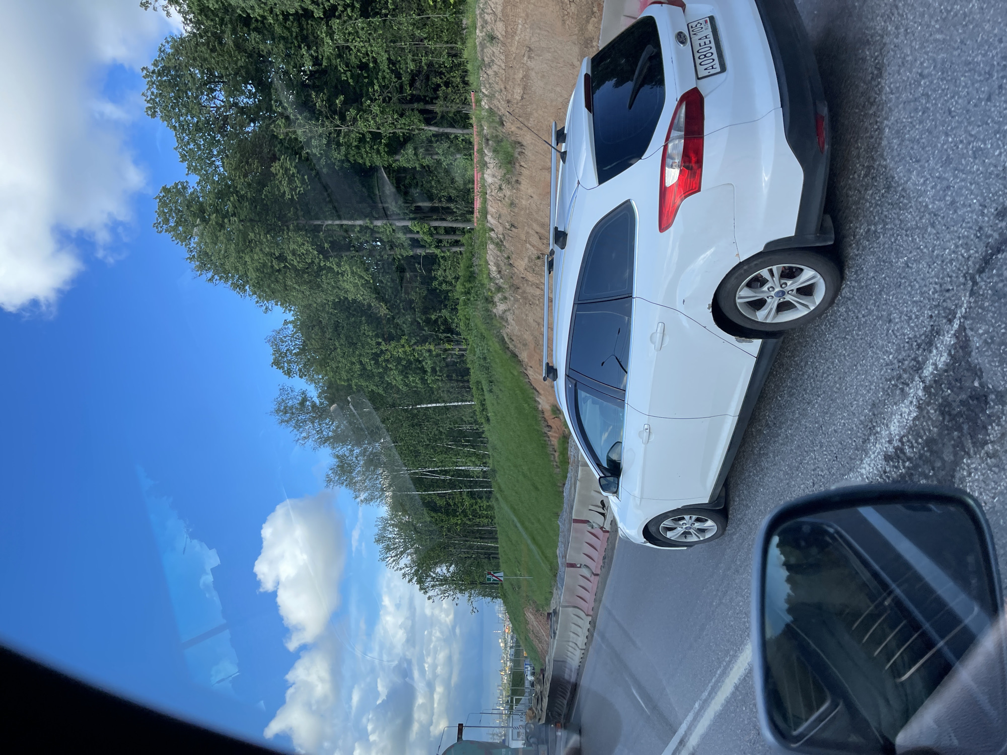 The image shows a view from a car traveling on a road in Novodrozhzhino, Russia. A white car is visible in the adjacent lane. The landscape includes trees, grass, and a blue sky with clouds. There are concrete barriers along the side of the road. The side mirror of the car taking the picture is also visible. The number plate of the white car is A080EA 105 RUS.