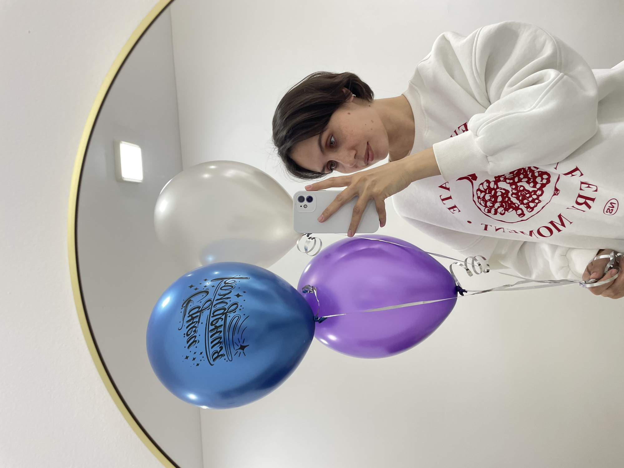 The image depicts a mirror selfie taken indoors, showcasing a person holding balloons. The central subject, appearing to be a young individual with short dark hair, is reflected in a large, round mirror framed with a thin gold rim. They are wearing a white oversized sweatshirt that features a mirrored red pomegranate design and stylized text. The person holds a light-colored smartphone in what appears to be their left hand (due to the mirror's reflection), aiming it to capture the reflection of themselves and the balloons.
Three balloons are prominently displayed: a metallic blue balloon, a purple balloon, and a white balloon. The blue balloon is adorned with mirrored black text that reads "С Днём Рождения" (Happy Birthday in Russian), surrounded by star patterns. All balloons are tied with shiny silver ribbons, and the person appears to be holding the ribbons of at least two of them.
The background visible in the mirror's reflection consists of plain white walls. A bright, square light fixture is also reflected in the upper left portion of the mirror. The lighting is bright and even, typical of an indoor setting. The person's expression is one of concentration, as they look towards the phone's screen while capturing the image. The presence of balloons, particularly the one with a "Happy Birthday" message, suggests a celebration or festive occasion.