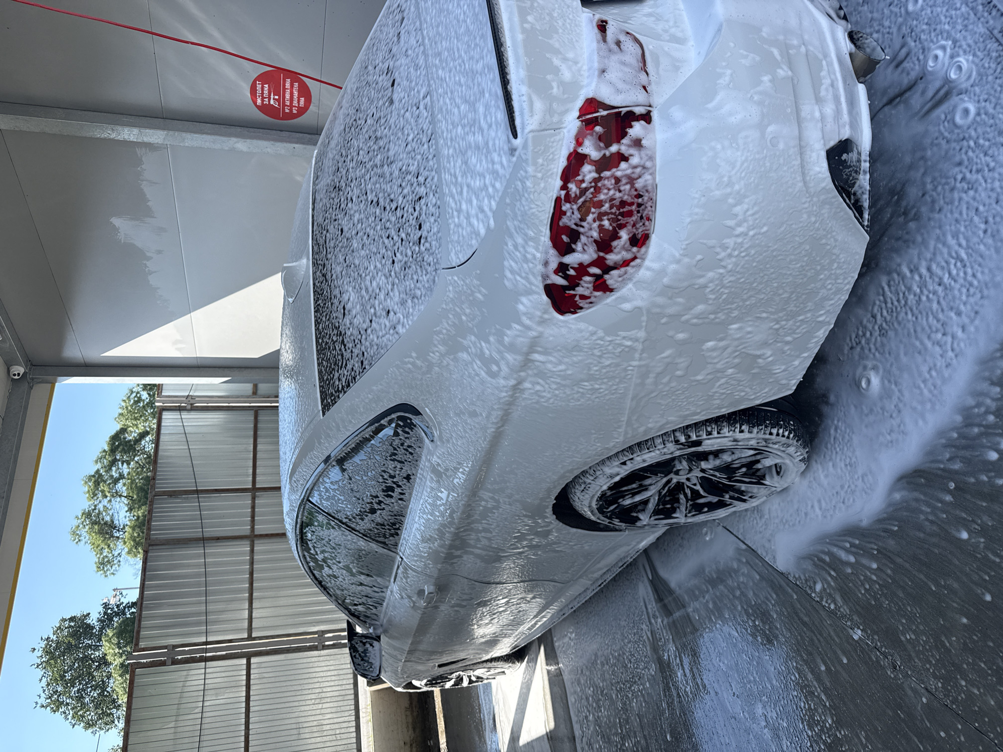 A white BMW coupe is parked at a self-service car wash in Varna, Bulgaria, covered in a thick layer of white foam, indicating it is in the process of being washed. The car is seen from a rear three-quarter perspective, with its rear lights, wheels, and roofline visible through the suds. The car wash bay has a light gray concrete floor with a drain, and a corrugated metal fence surrounds the exterior. A red circular sign with Ukrainian text is affixed to the wall, indicating instructions for using the car wash equipment. The sky is clear and blue, suggesting a sunny day. The scene captures a moment of routine car maintenance in an urban outdoor setting.