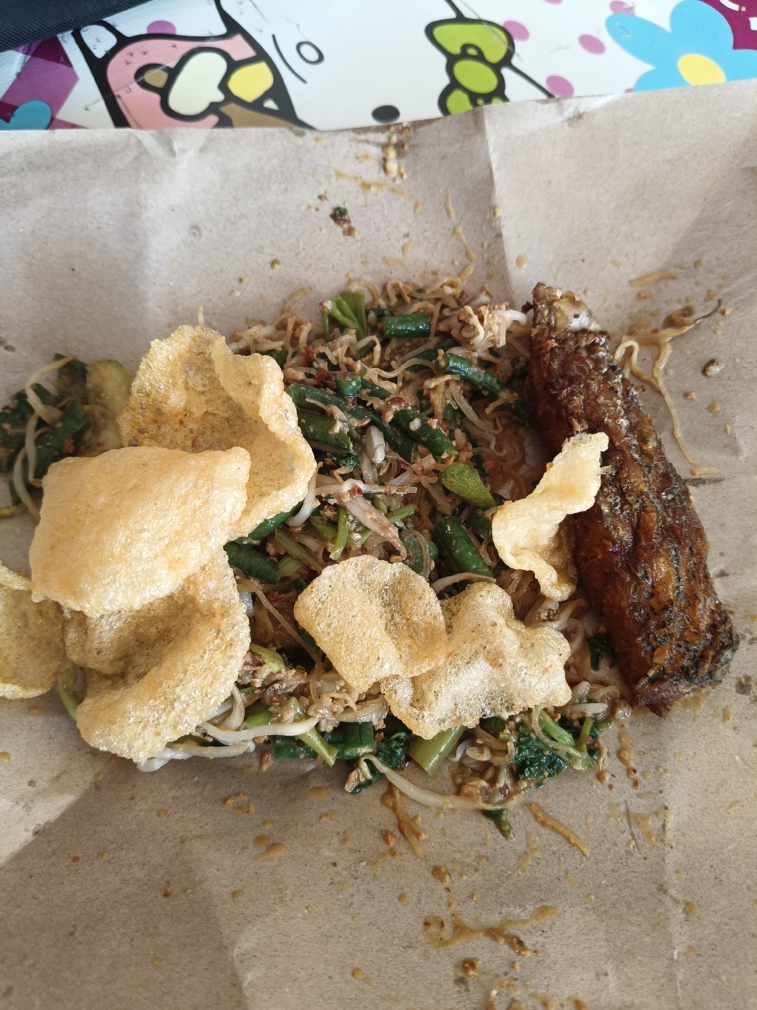 The image shows a close-up view of a dish of mixed vegetables and a piece of fried fish, served on brown paper. The dish appears to be a type of Indonesian salad, possibly "Gado-gado" or "Pecel," with ingredients such as chopped green beans, bean sprouts, and a peanut-based sauce. Several crispy crackers, likely krupuk, are placed on top of the salad. The fried fish is positioned to the right of the salad. The overall presentation is casual, with the food directly on the paper, suggesting it might be street food or a simple meal. The background shows a colorful, patterned surface, possibly a tablecloth or placemat, with a cartoon-like character design. There are no people visible in the image. The lighting suggests it is daytime, possibly outdoors or near a window. The scene is static, focusing entirely on the food. The location in Rancaseneng, Indonesia, is not visually evident from the provided image itself, but this context has been provided as part of the prompt.