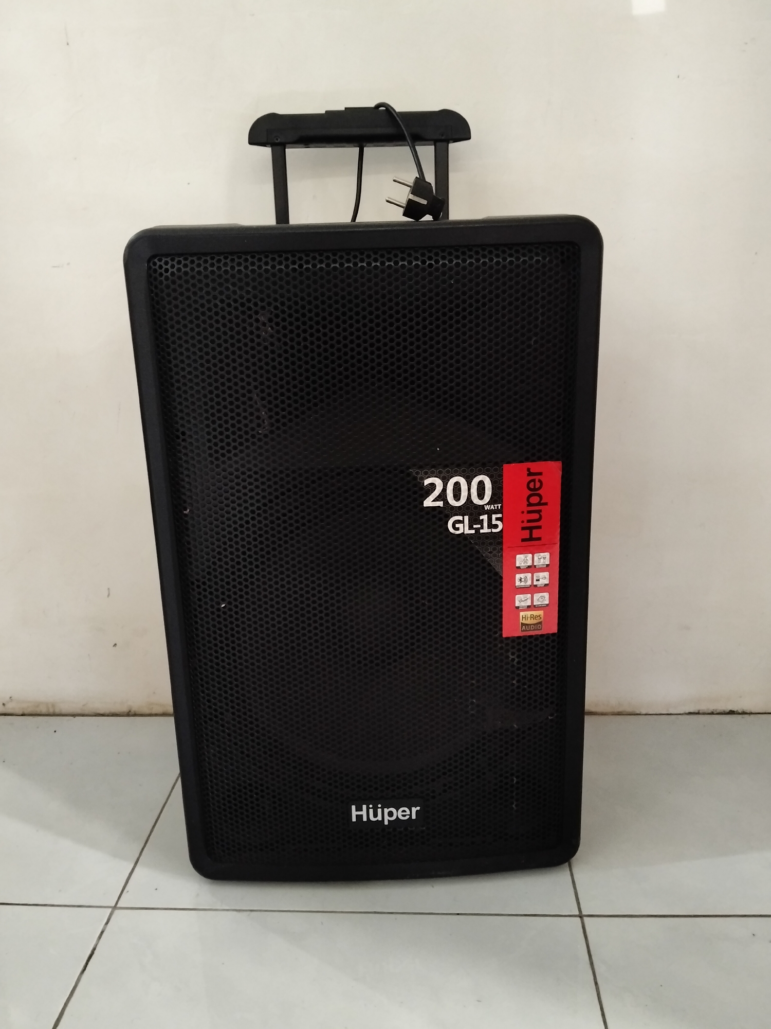 A black powered speaker with a handle and wheels is placed on a tiled floor against a white wall. The speaker has a prominent mesh grille and branding that reads "200 WATT GL-15" and "Hüper" with a red sticker. The sticker contains several icons and the text "Hi-Res AUDIO". A power cord with a plug is visible at the top of the speaker. The environment appears to be indoors, likely a residential or storage space, in Tasikmadu, Indonesia.